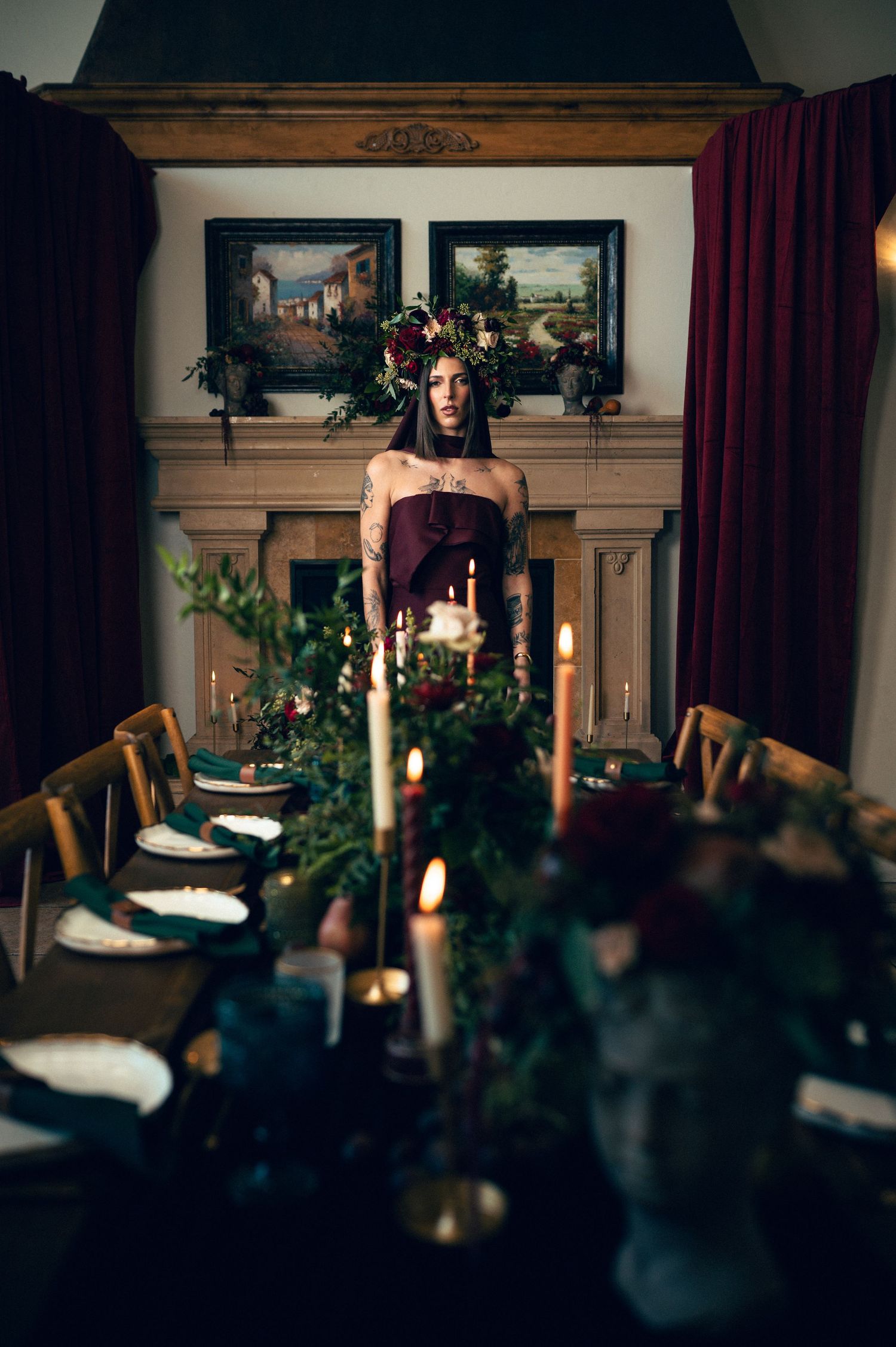 Woman in burgundy gown with floral crown stands at candlelit gothic dining table with lush greenery centerpiece.