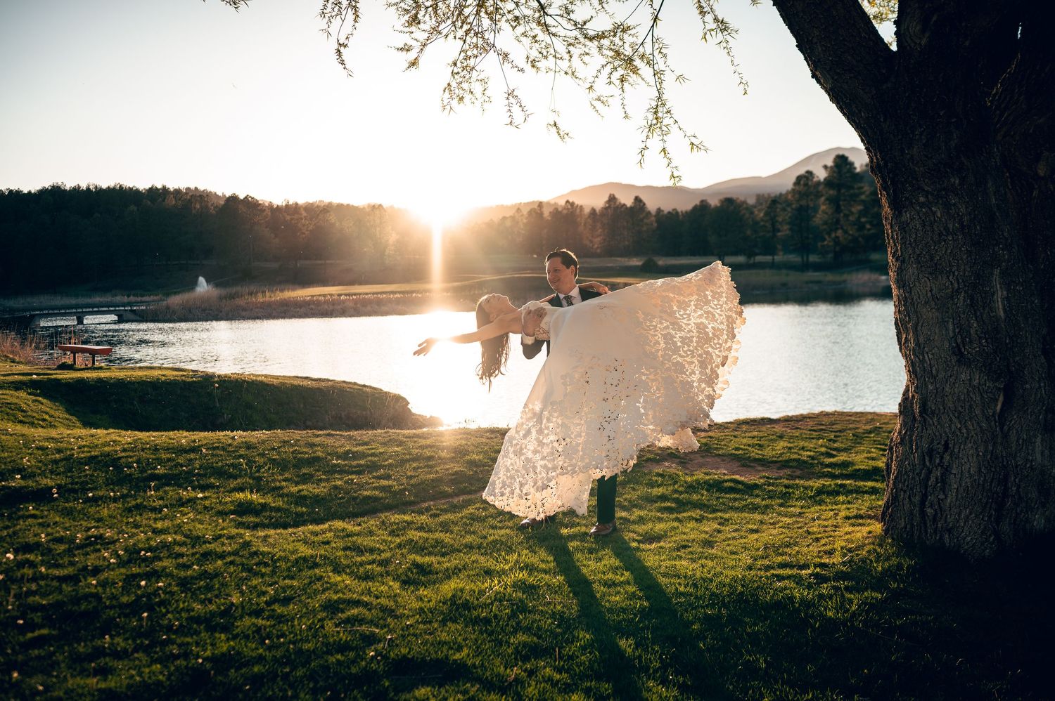 Groom lifting bride in lace wedding dress at golden sunset by a lakeside tree on green grass.