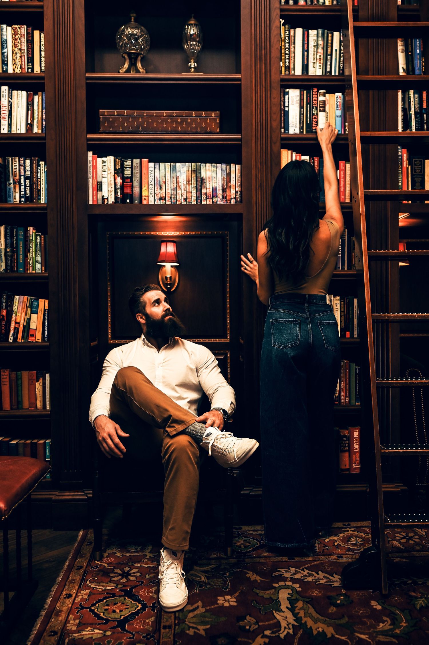 A bearded man sits in a leather chair while a woman reaches for books on a tall shelf in a dark library.