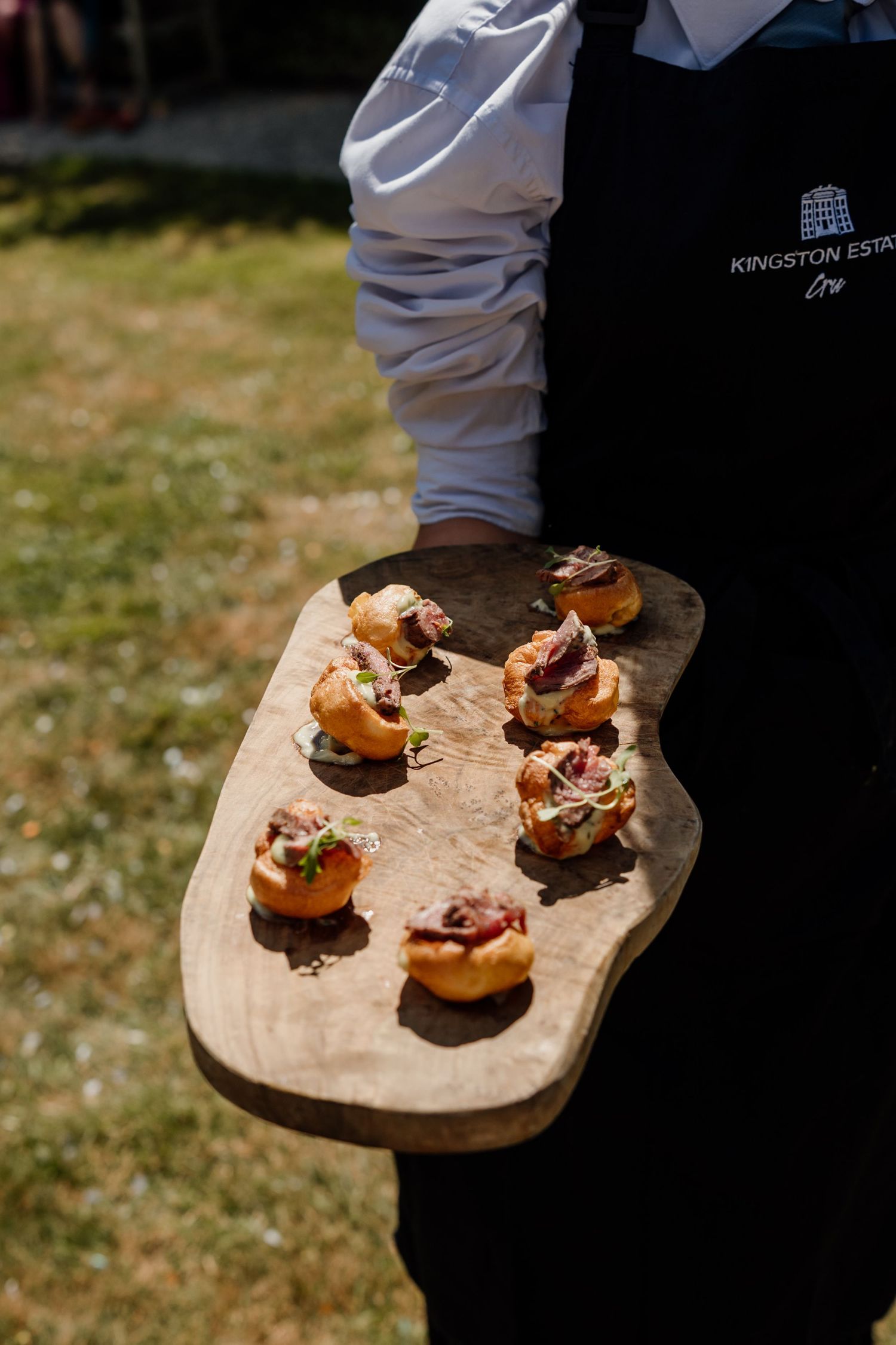 A Kingston Estate server holds a rustic wooden board with elegant bite-sized canapes at an outdoor catering event.