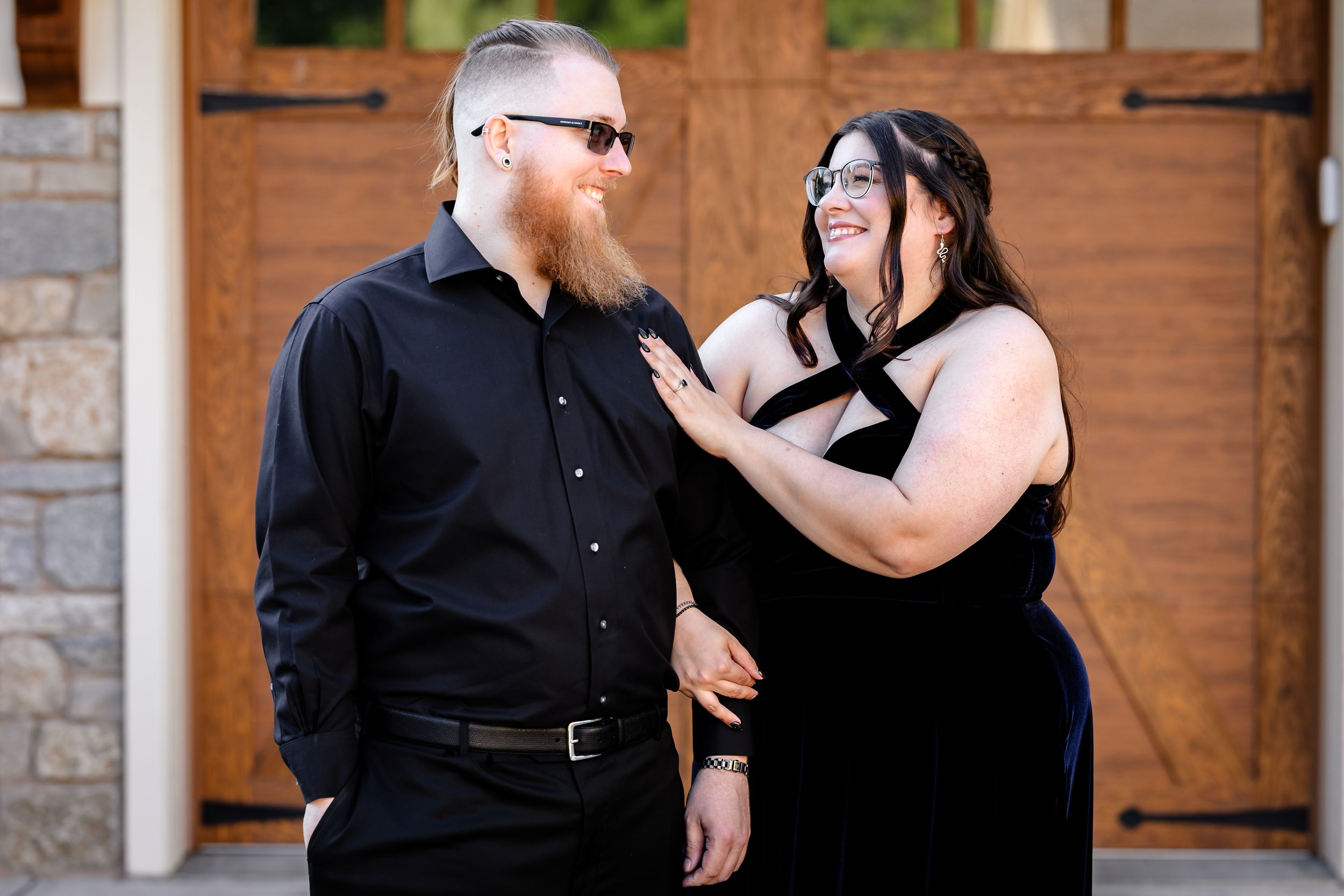 Couple in matching black outfits smiling at each other outside rustic barn doors during engagement photos.