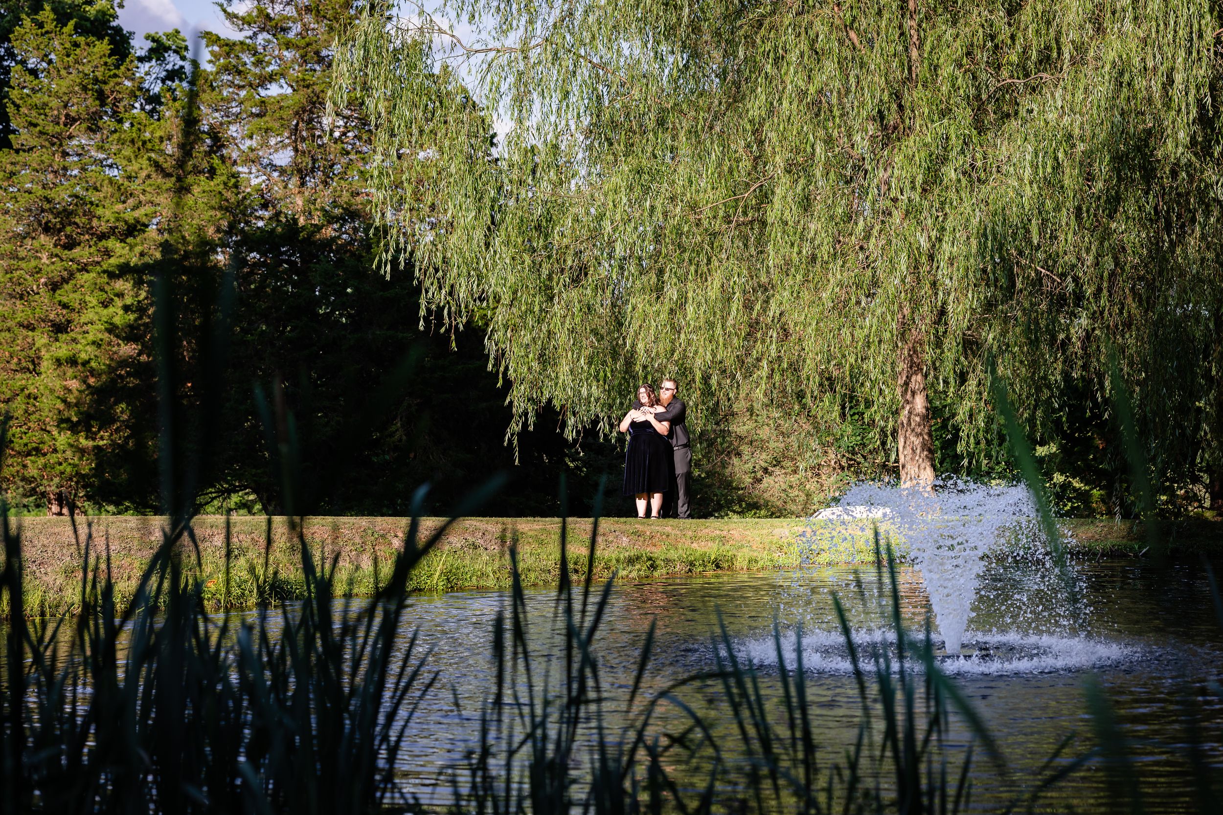 Person standing near a decorative pond fountain surrounded by weeping willow trees in a lush park.
