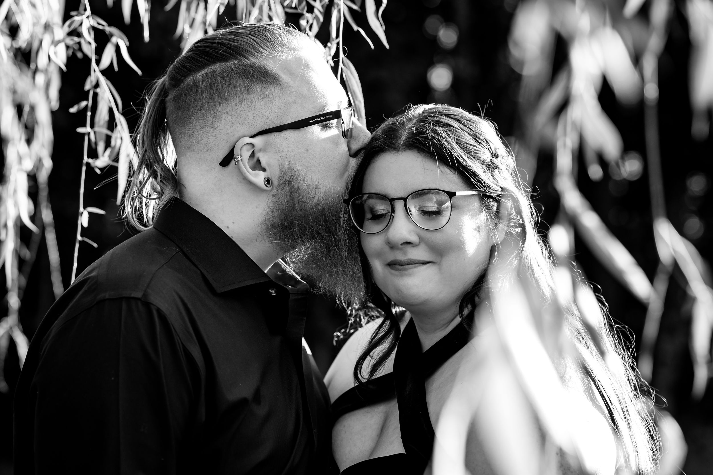 Black and white photo of a couple sharing a romantic moment, man kissing womans cheek outdoors.