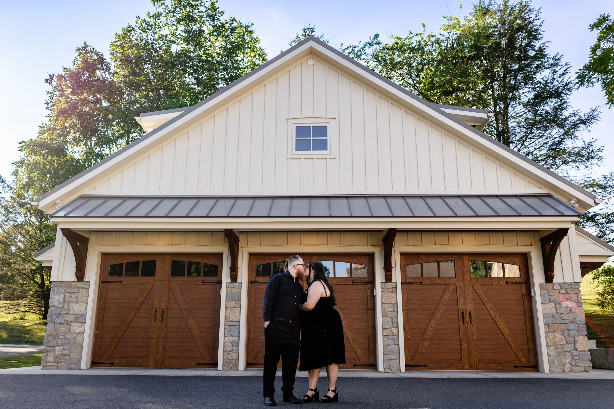 Couple sharing a romantic kiss in front of a charming three-car garage with wood doors and stone accents.
