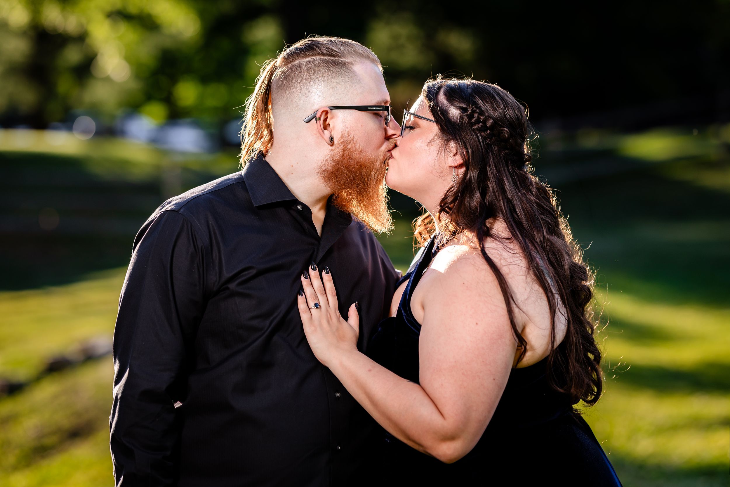 A couple shares a romantic kiss outdoors, dressed in black, with lush green grass and trees in the background.