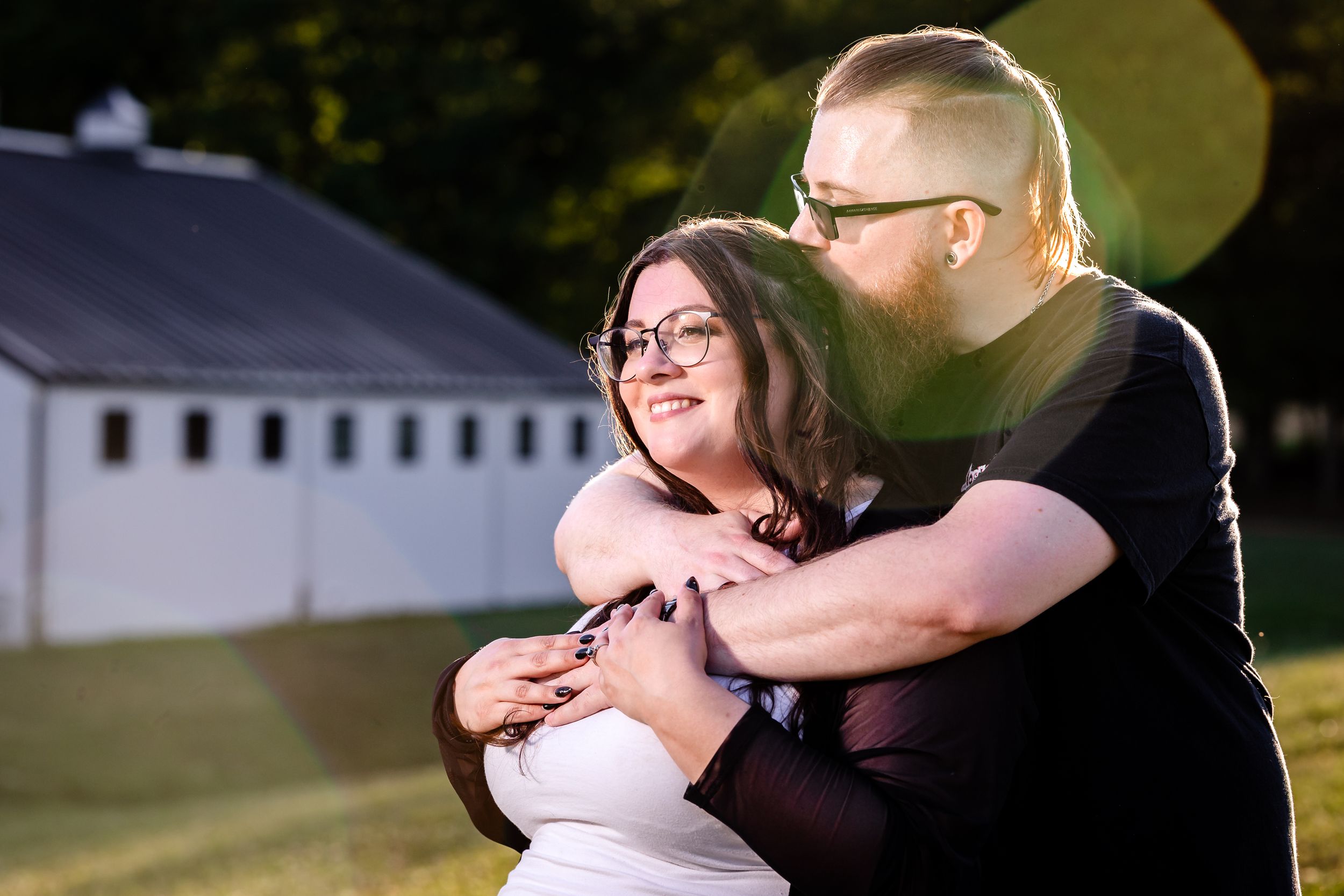 Couple embracing in golden hour sunlight near a white barn during outdoor engagement session.