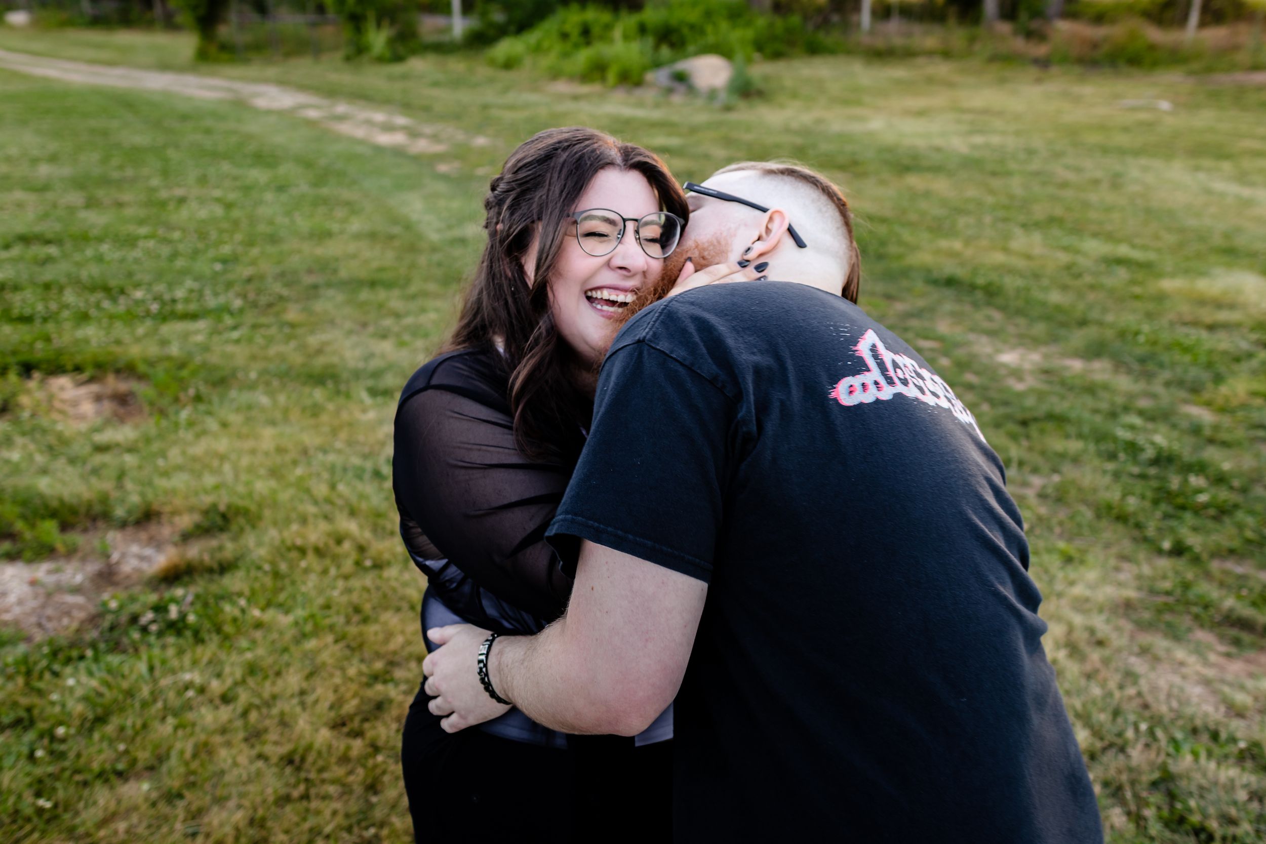 Woman laughing joyfully while being hugged by a man in a black t-shirt outdoors on green grass.