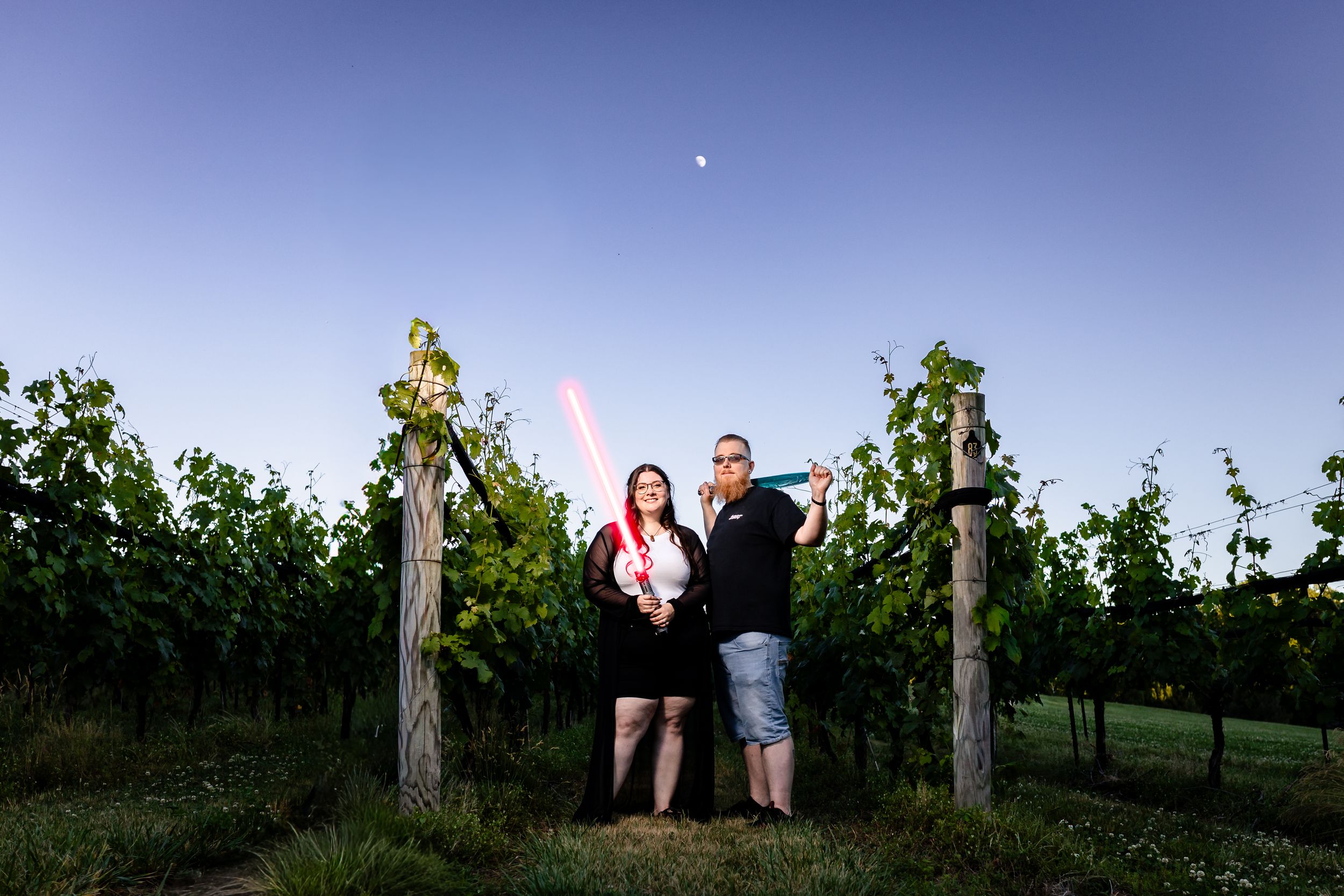 Couple posing with lightsabers in a lush vineyard at dusk with a crescent moon above.