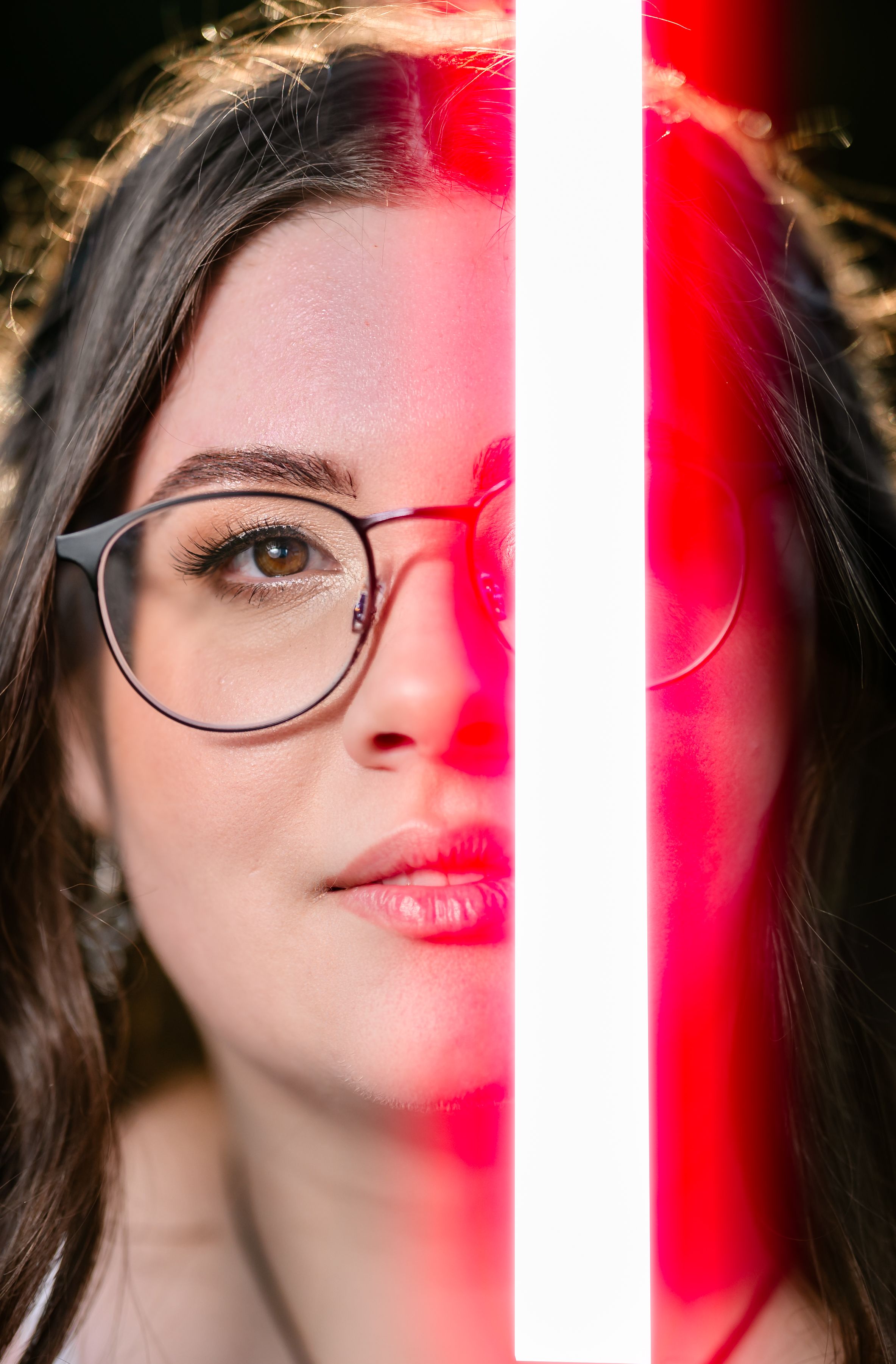Young woman with glasses poses behind a glowing red and white neon light in a striking portrait photo.
