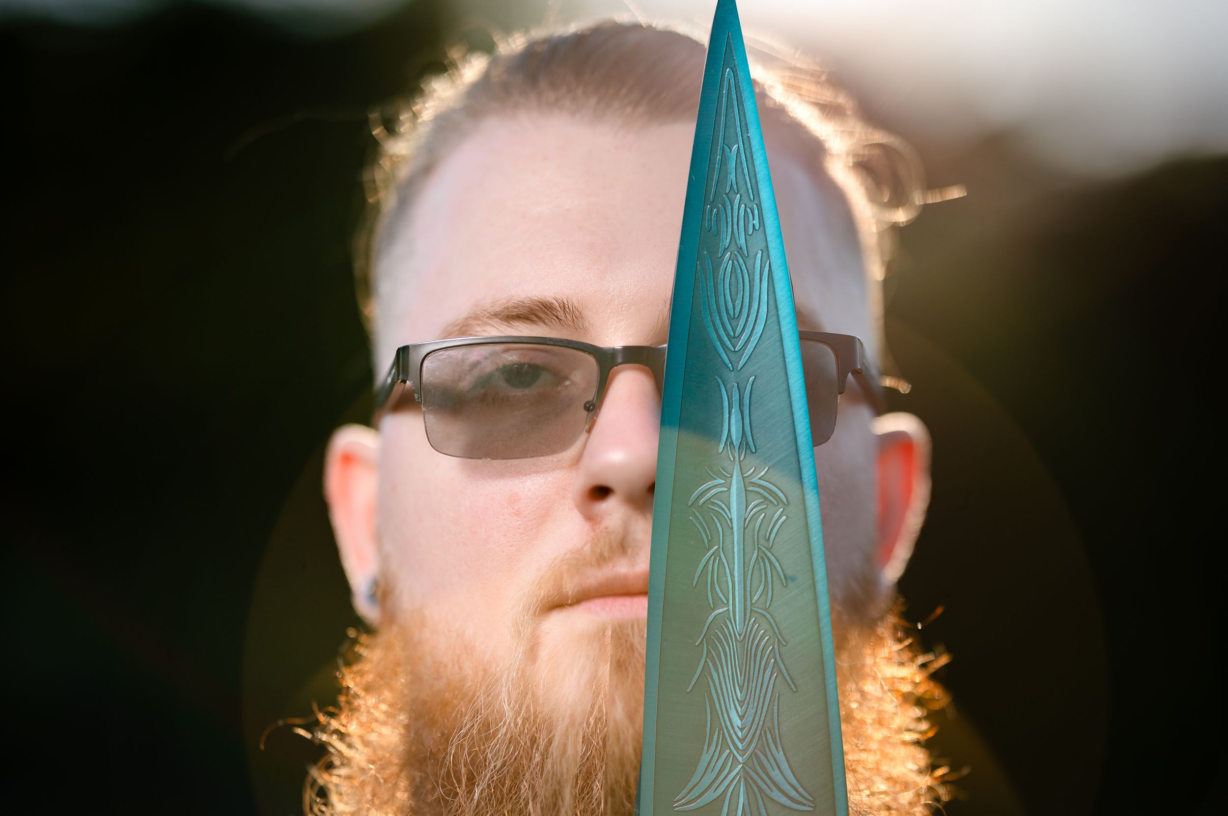 Bearded man with glasses peers behind a decorative teal blade in dramatic sunlit outdoor portrait.