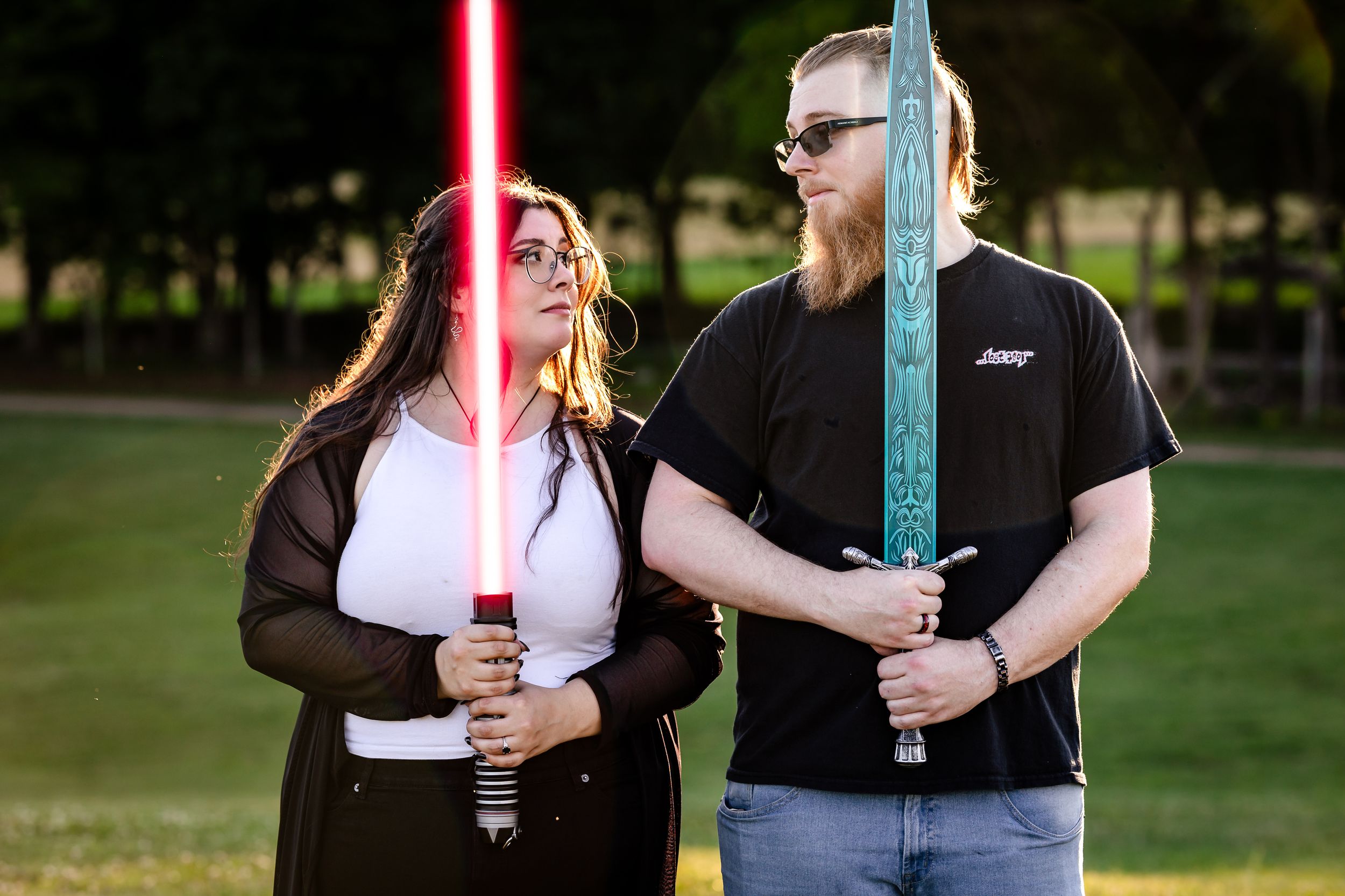 Couple posing with glowing red and yellow Star Wars lightsabers in a park at dusk.