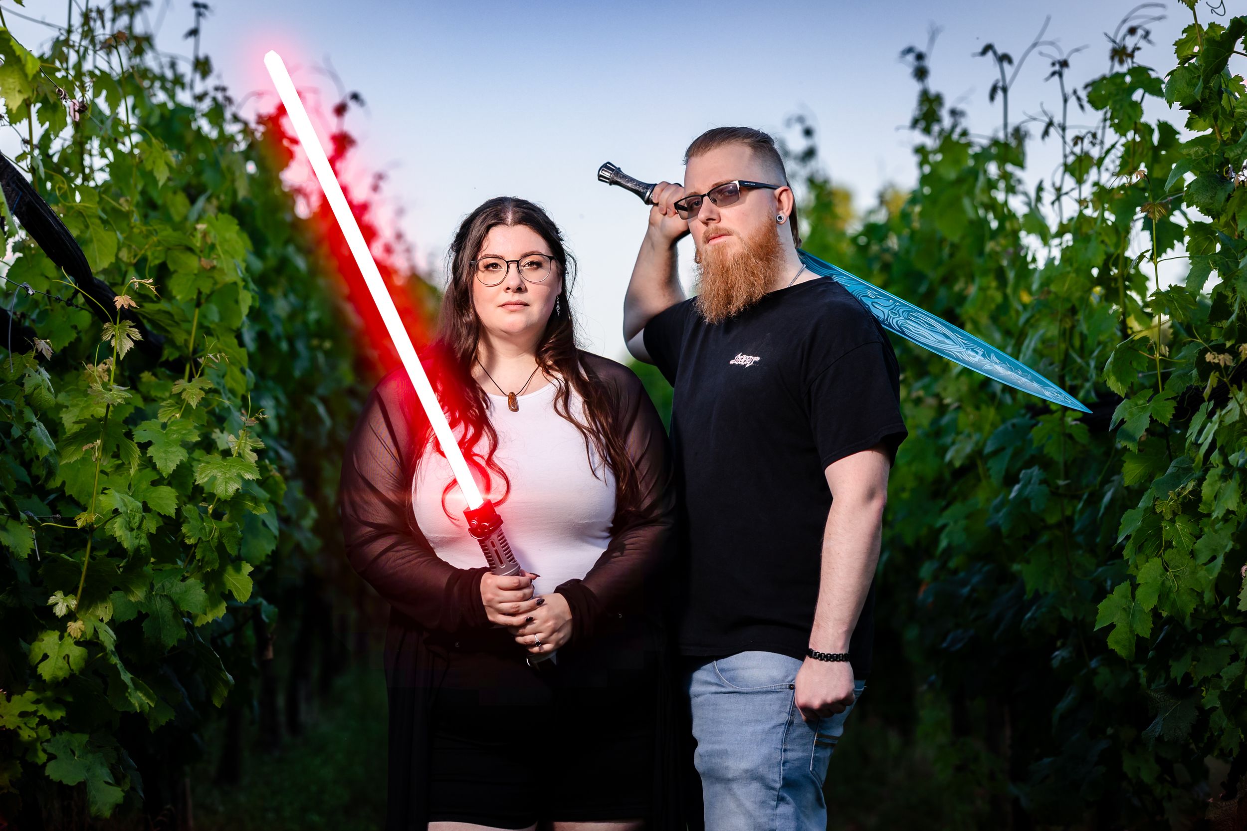 Two Star Wars fans pose with glowing lightsabers in a vineyard at dusk, woman holds red saber, man holds blue.