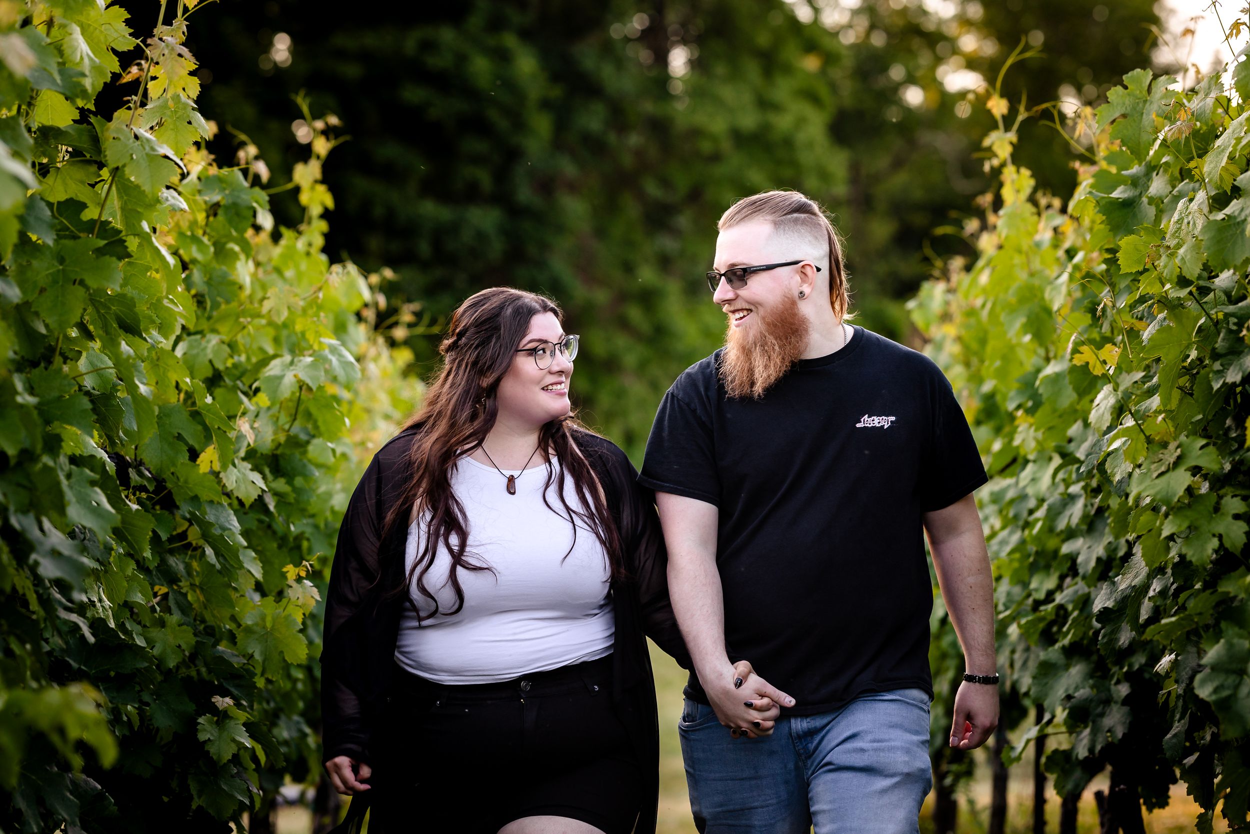 Smiling couple holding hands and gazing at each other between green vineyard rows.