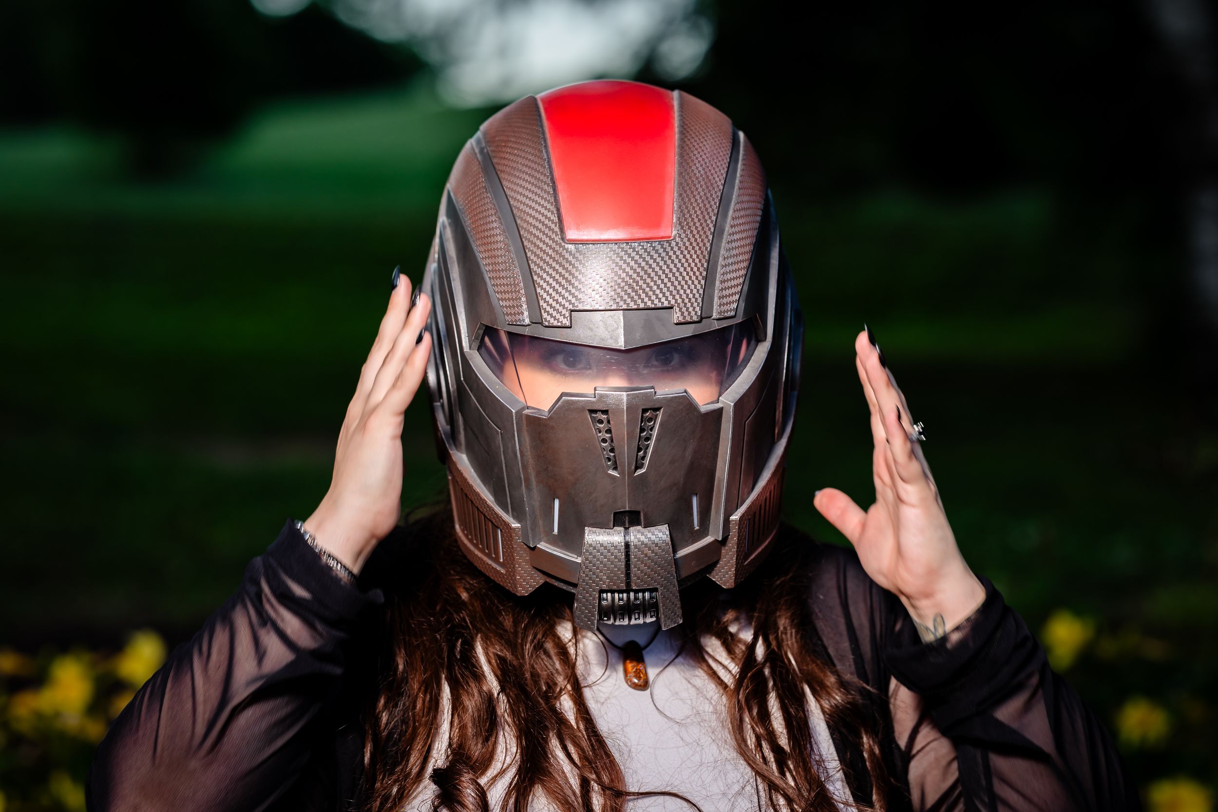 Woman wearing a futuristic gray and red N7 Mass Effect helmet outdoors in a garden setting.