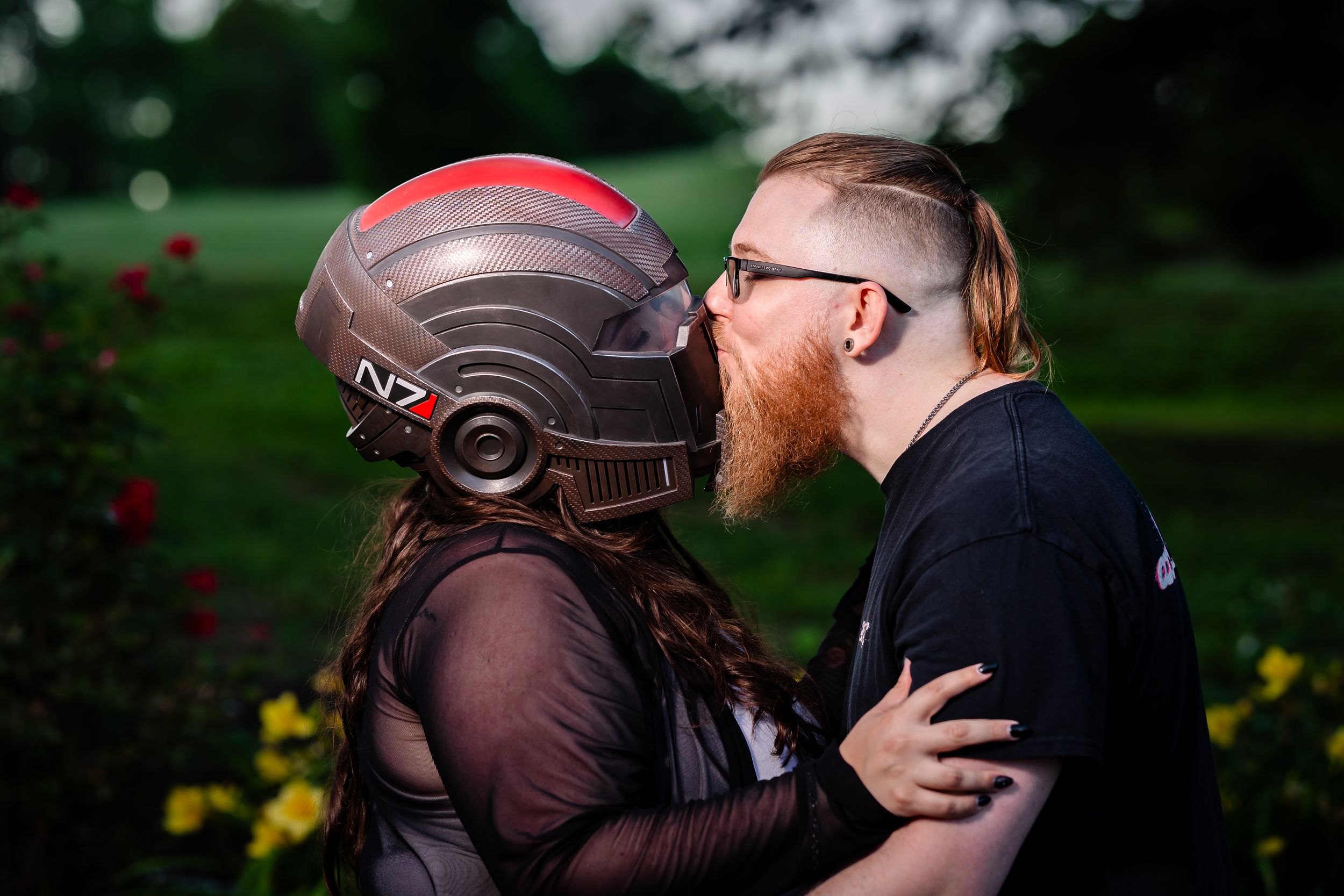 Bearded man kissing a woman wearing an N7 Mass Effect helmet in a lush garden setting.