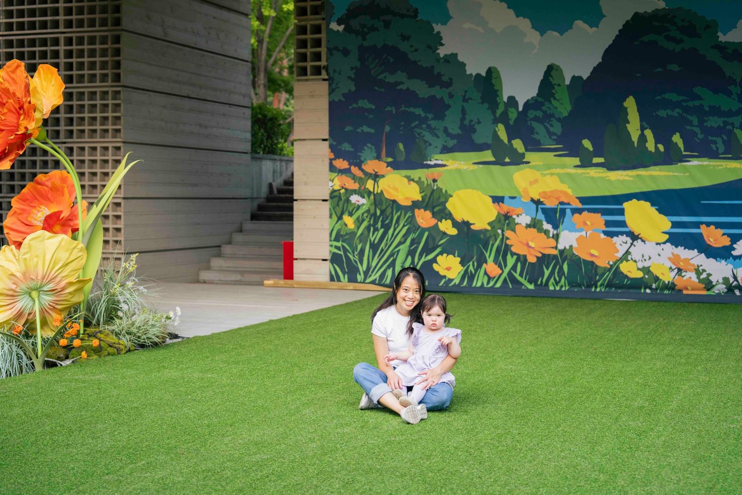 Woman and child sitting on artificial grass in front of a colorful floral mural with orange poppies outdoors.