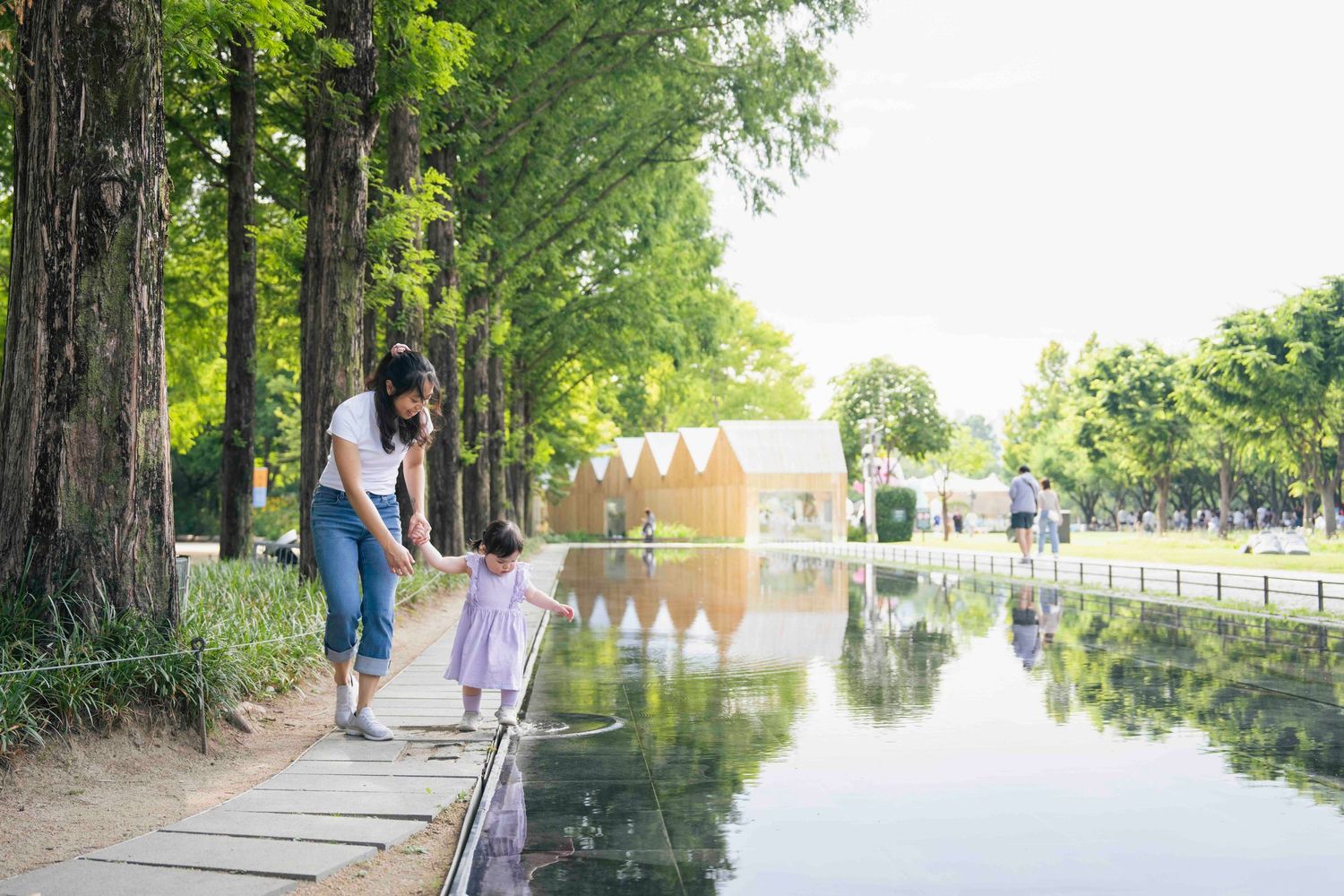 Mother and child walking along a serene park pond path lined with tall green trees on a sunny day.