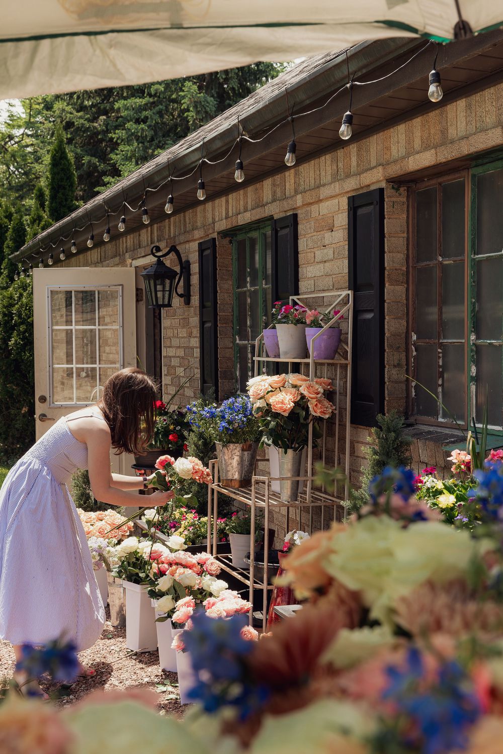 Woman in white dress arranging colorful flowers outside a charming brick flower shop with string lights.