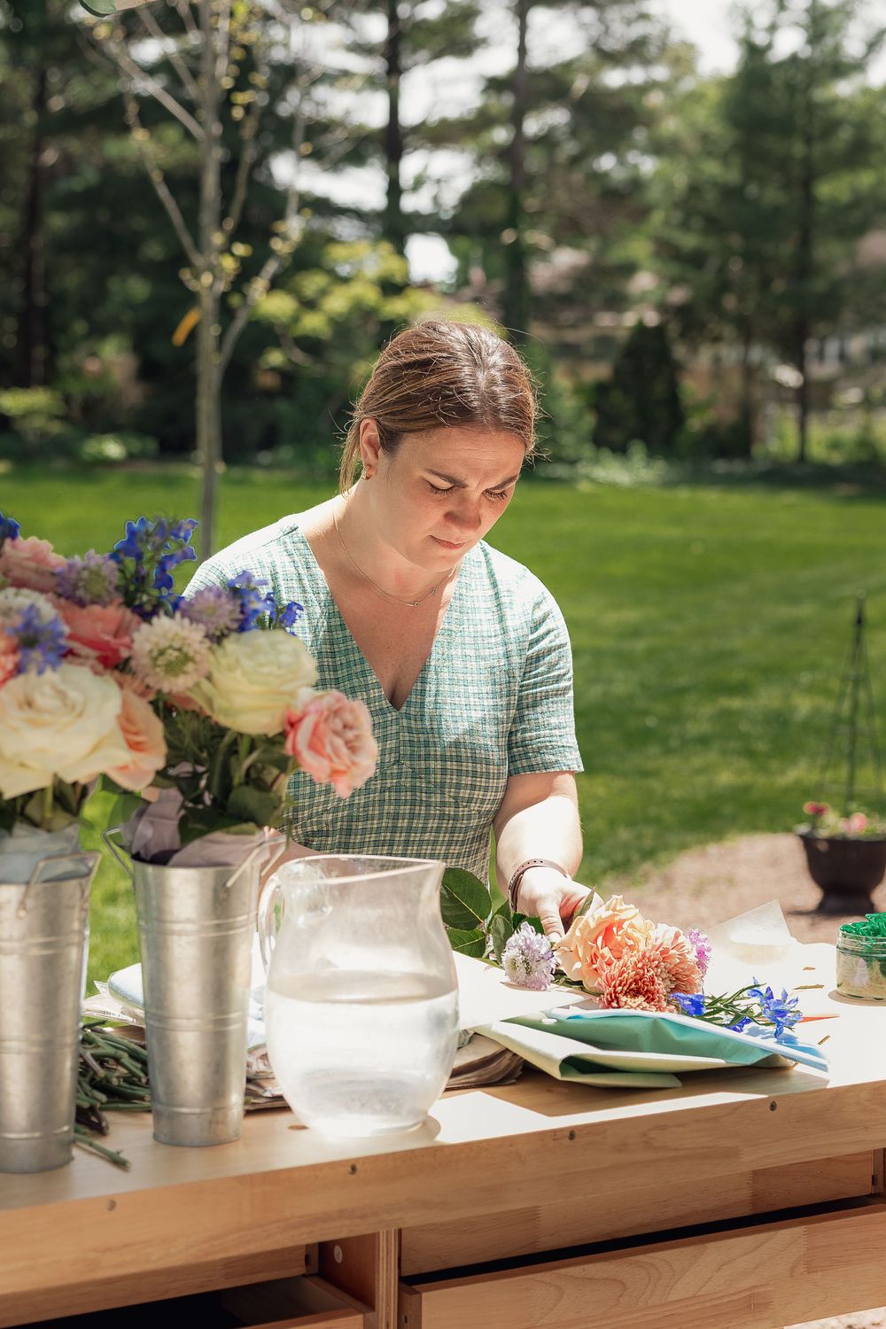 Woman arranging fresh flowers at an outdoor wooden table surrounded by lush greenery.