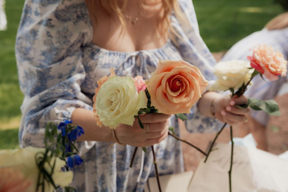 Women in floral dress holding cream and peach roses at an outdoor garden event.