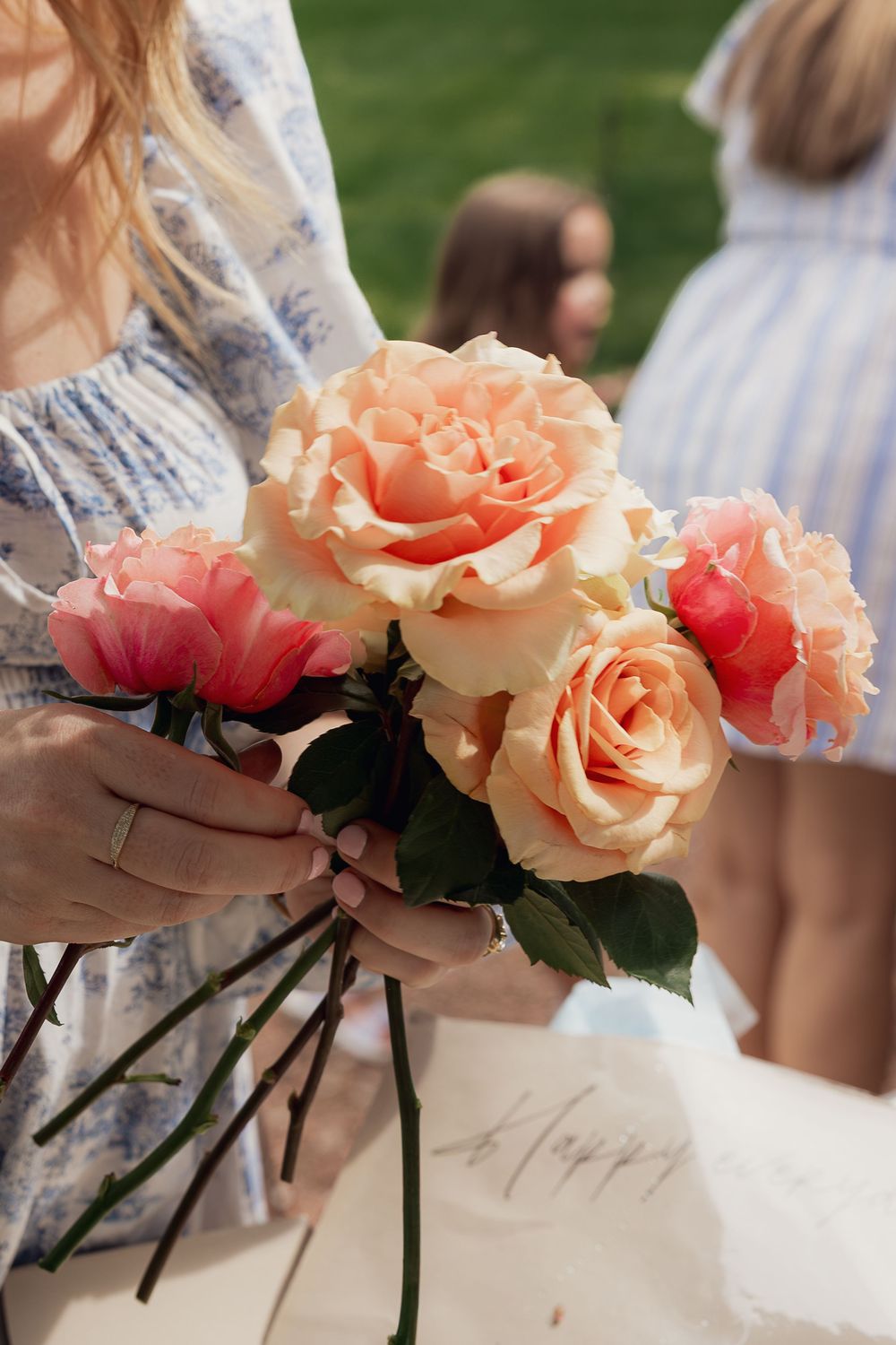 Woman in floral dress holding a bouquet of peach and pink roses at an outdoor celebration.