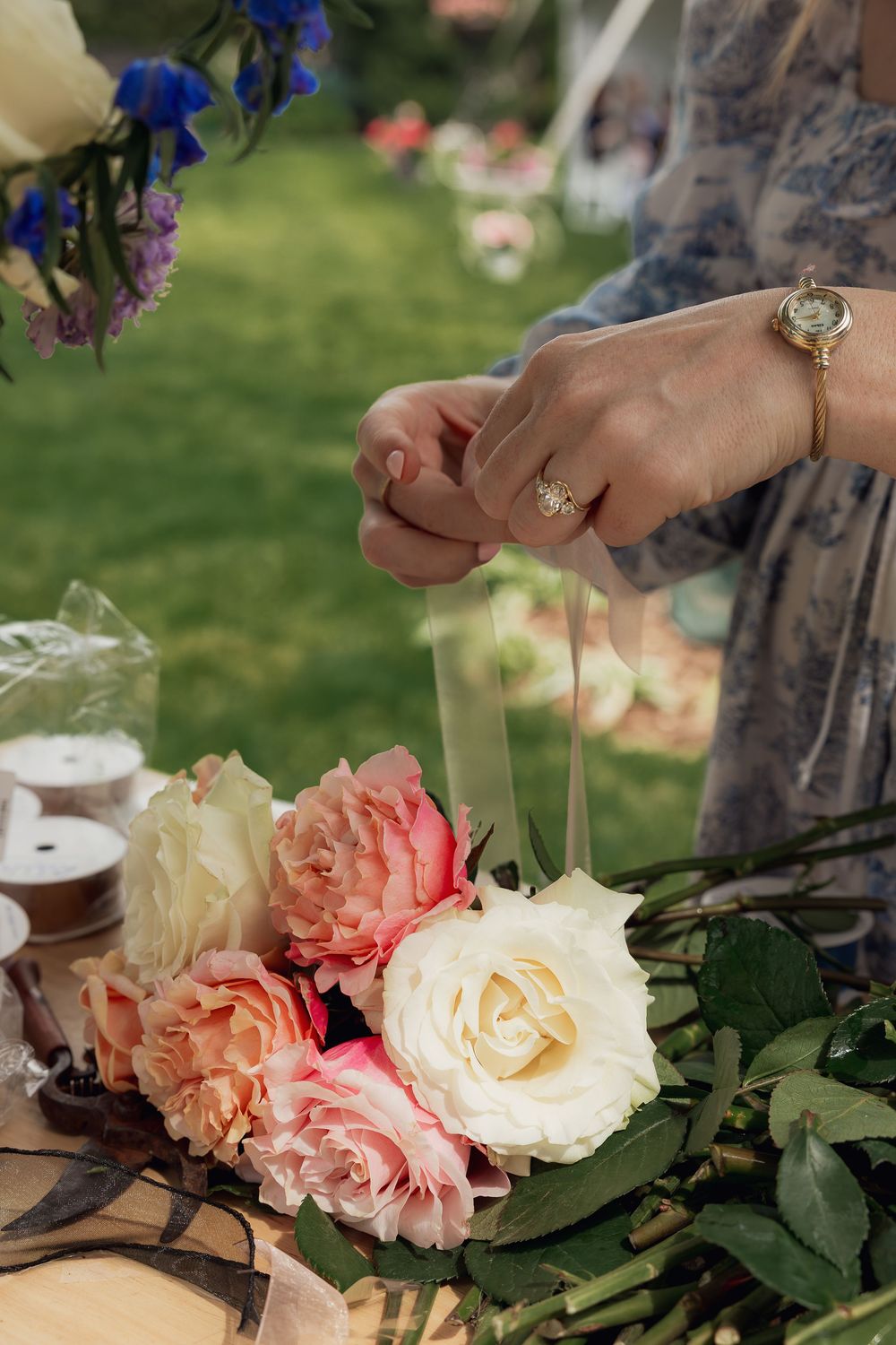 Florist arranging pink and cream roses with ribbon at outdoor wedding flower workshop on green grass background.