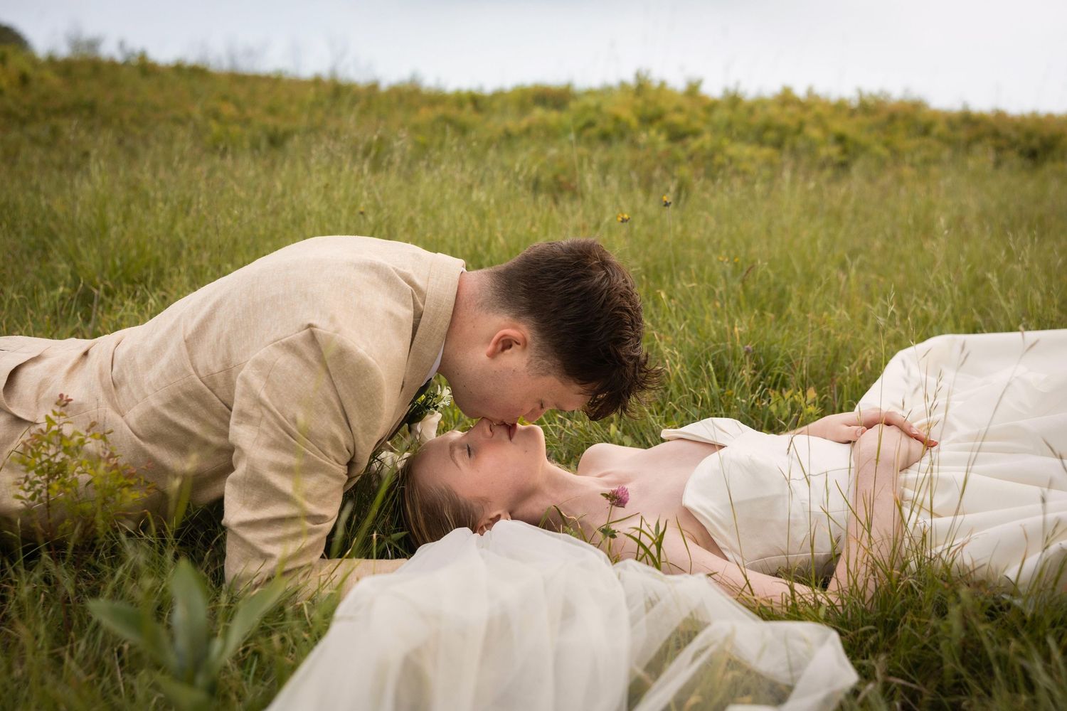 Romantic couple sharing a kiss while lying in a lush green meadow, dressed in elegant wedding attire.