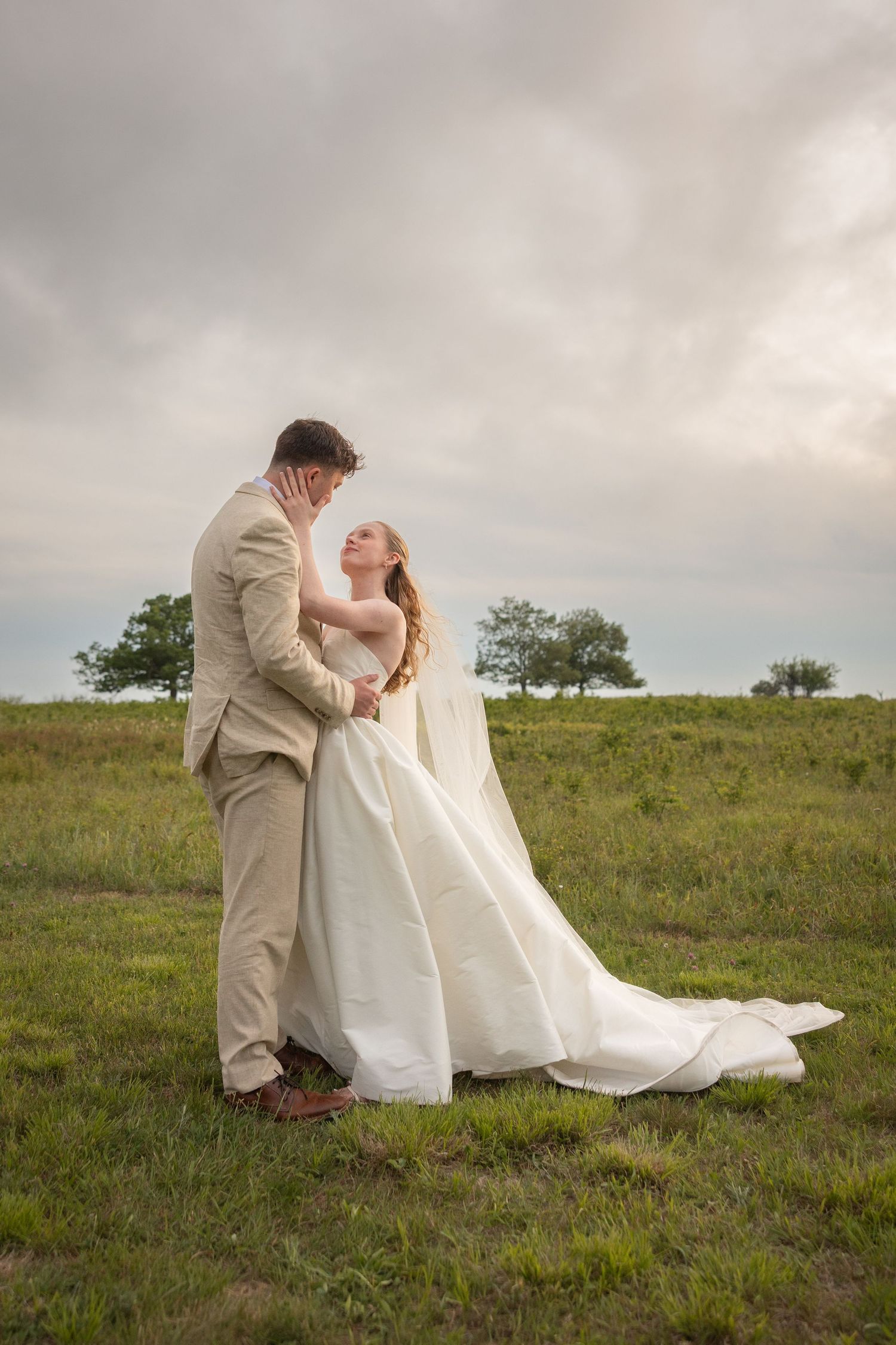 Bride in white gown and groom in tan suit embrace in a green field under cloudy skies.