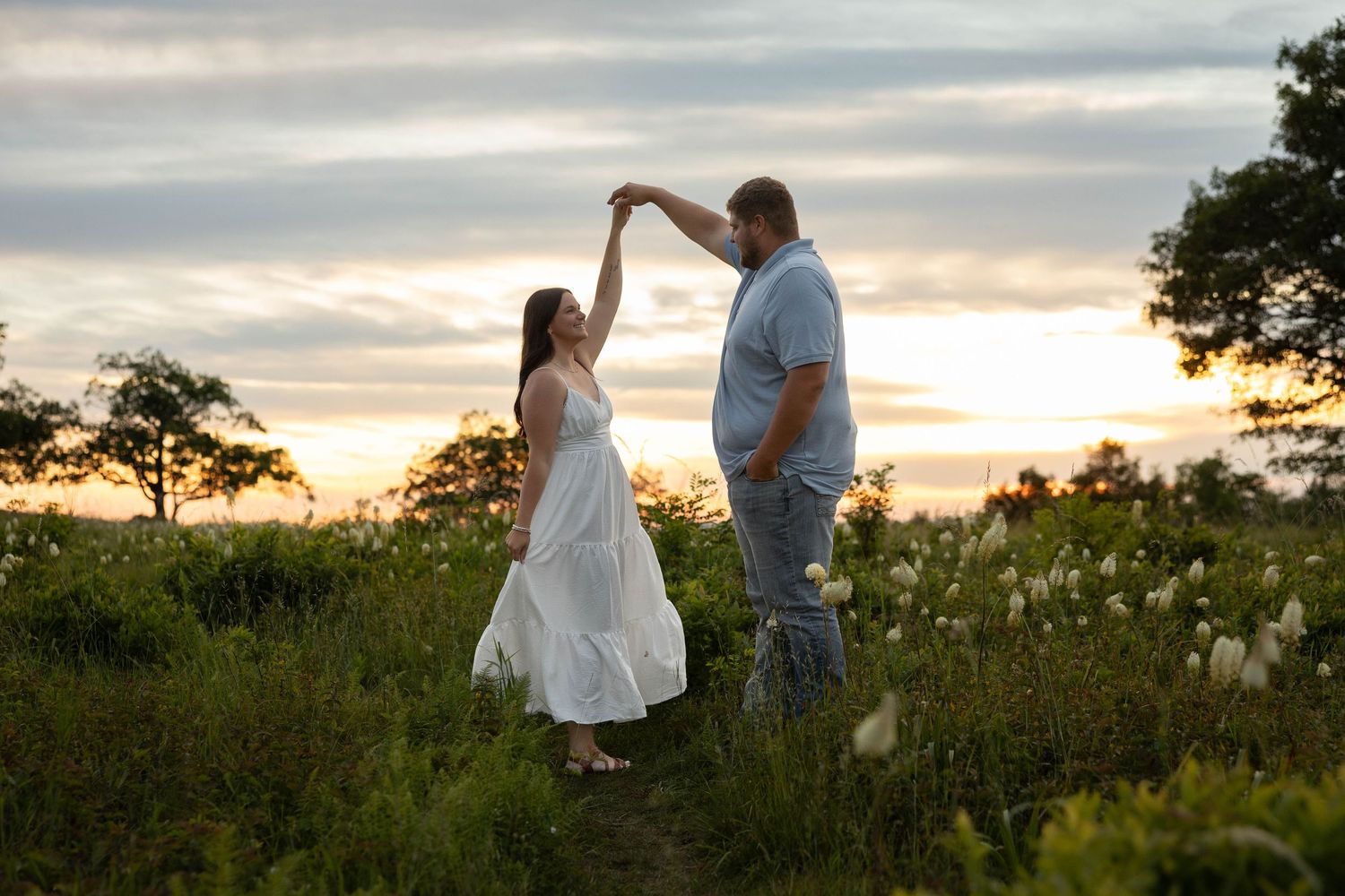 Couple dancing at sunset in a wildflower field, man spinning woman in white dress, romantic golden hour outdoor photo.