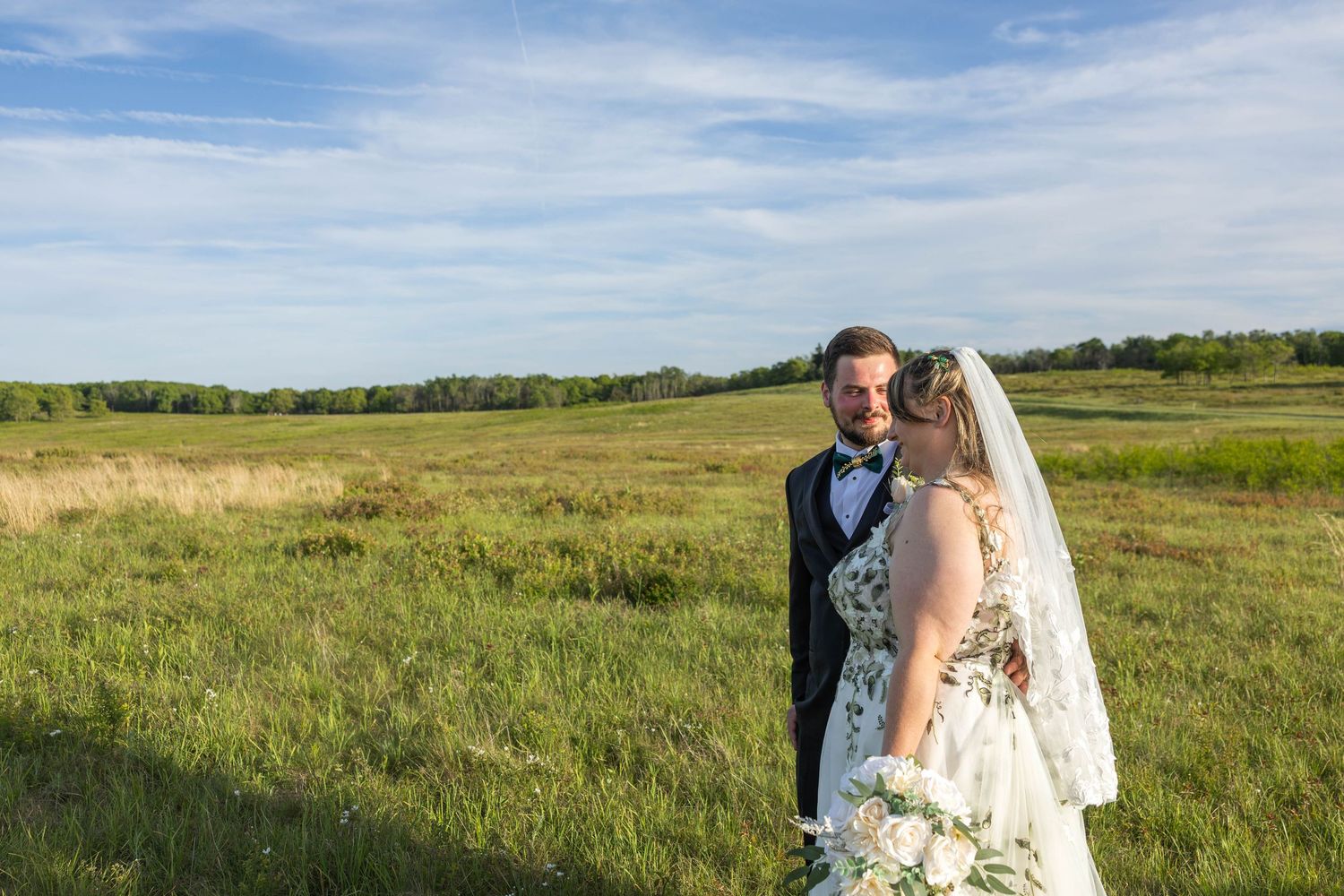 Bride and groom stand in a lush green field under a blue sky on their wedding day.