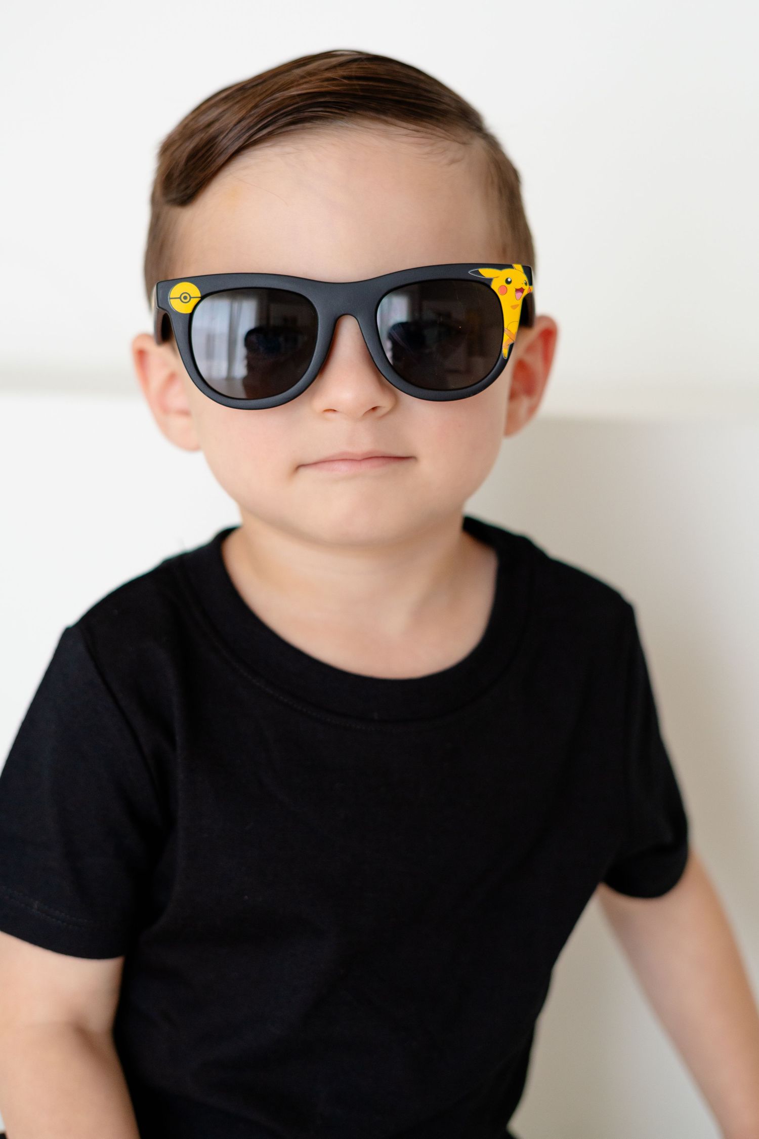 Young boy wearing black t-shirt and yellow giraffe print sunglasses posing confidently against white background.
