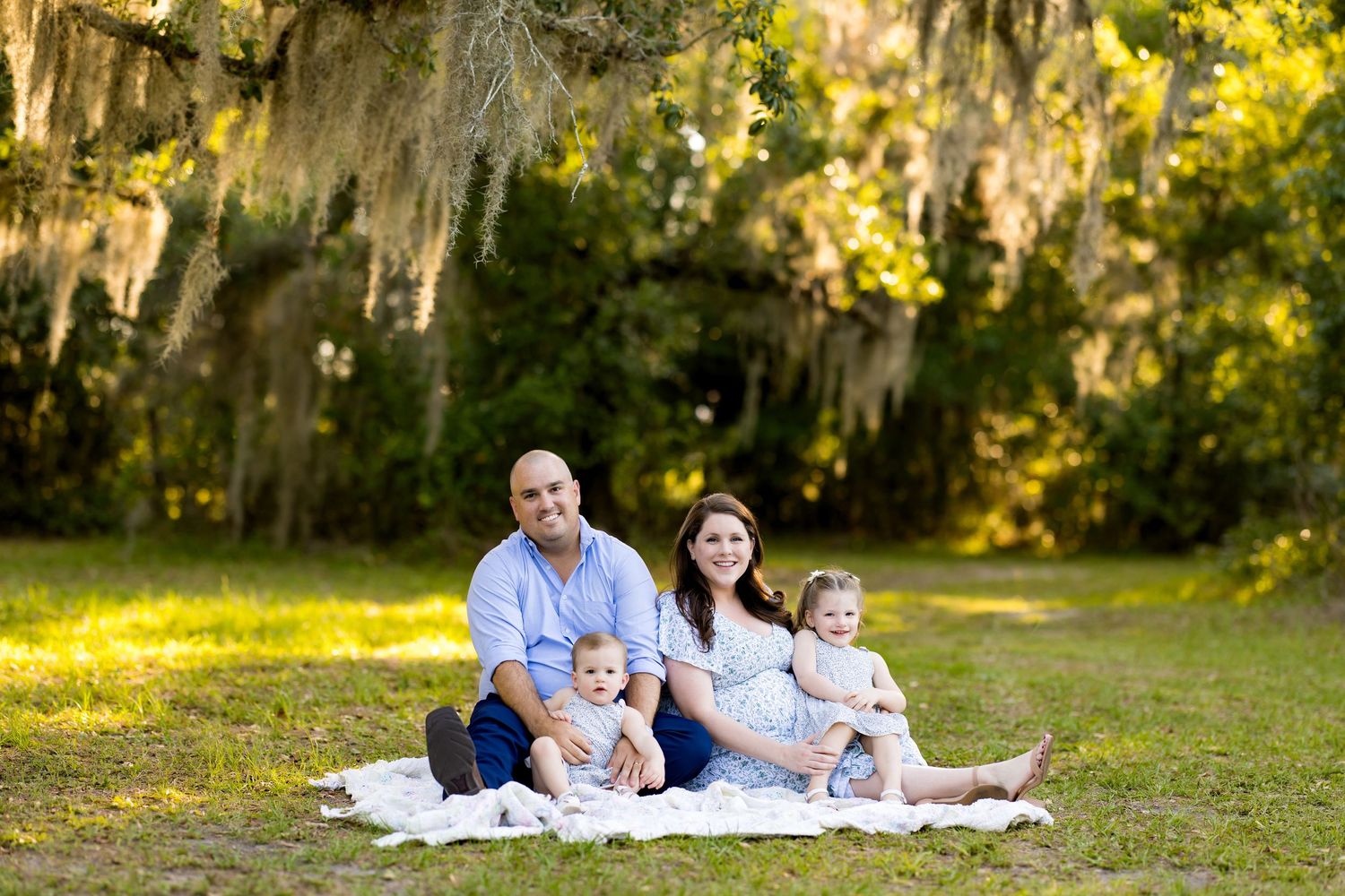 Family of four sitting on blanket in sunny park under Spanish moss-draped trees for outdoor portrait.