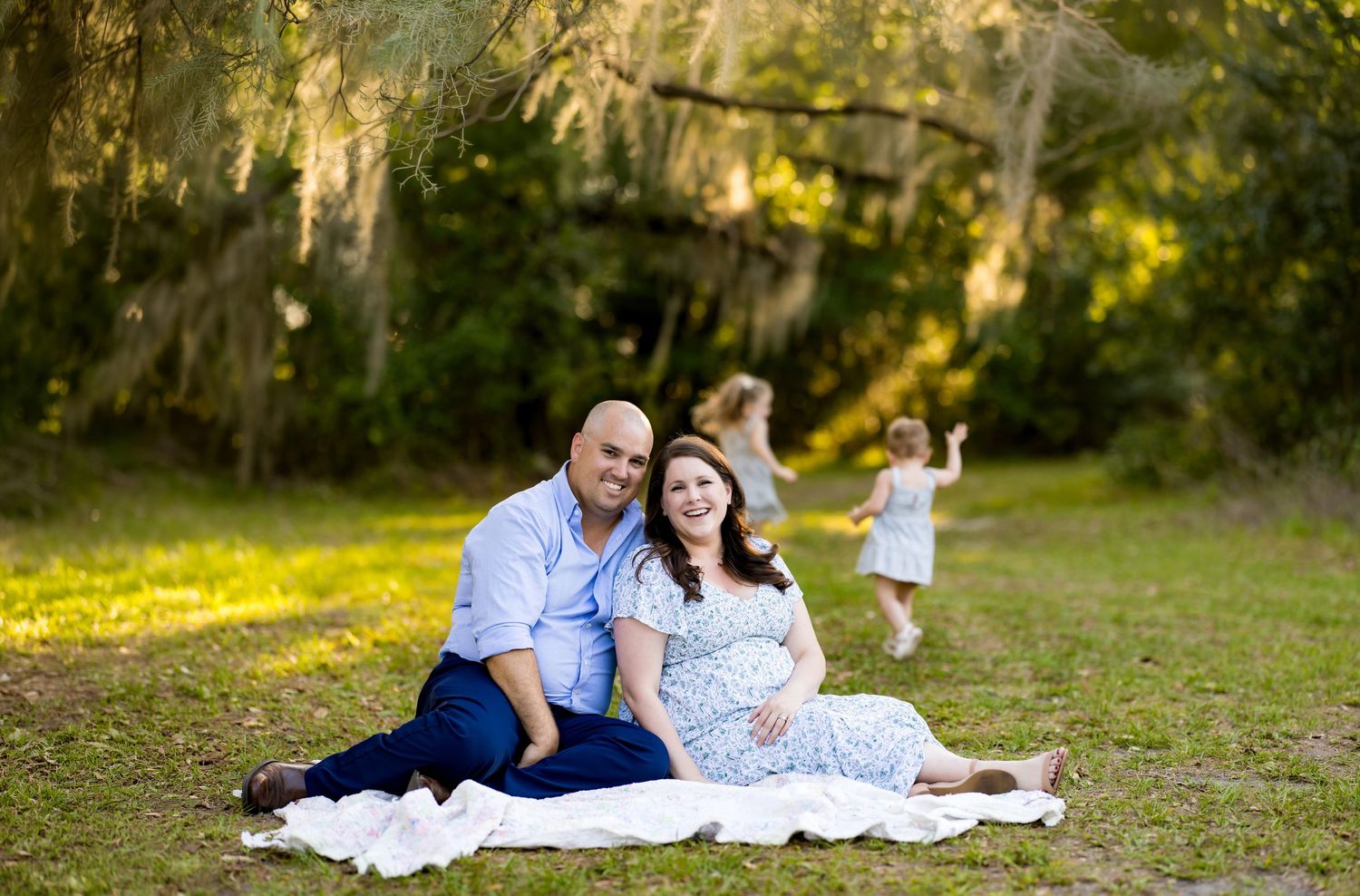 Pregnant couple sits on blanket outdoors smiling, children playing behind them among mossy trees.