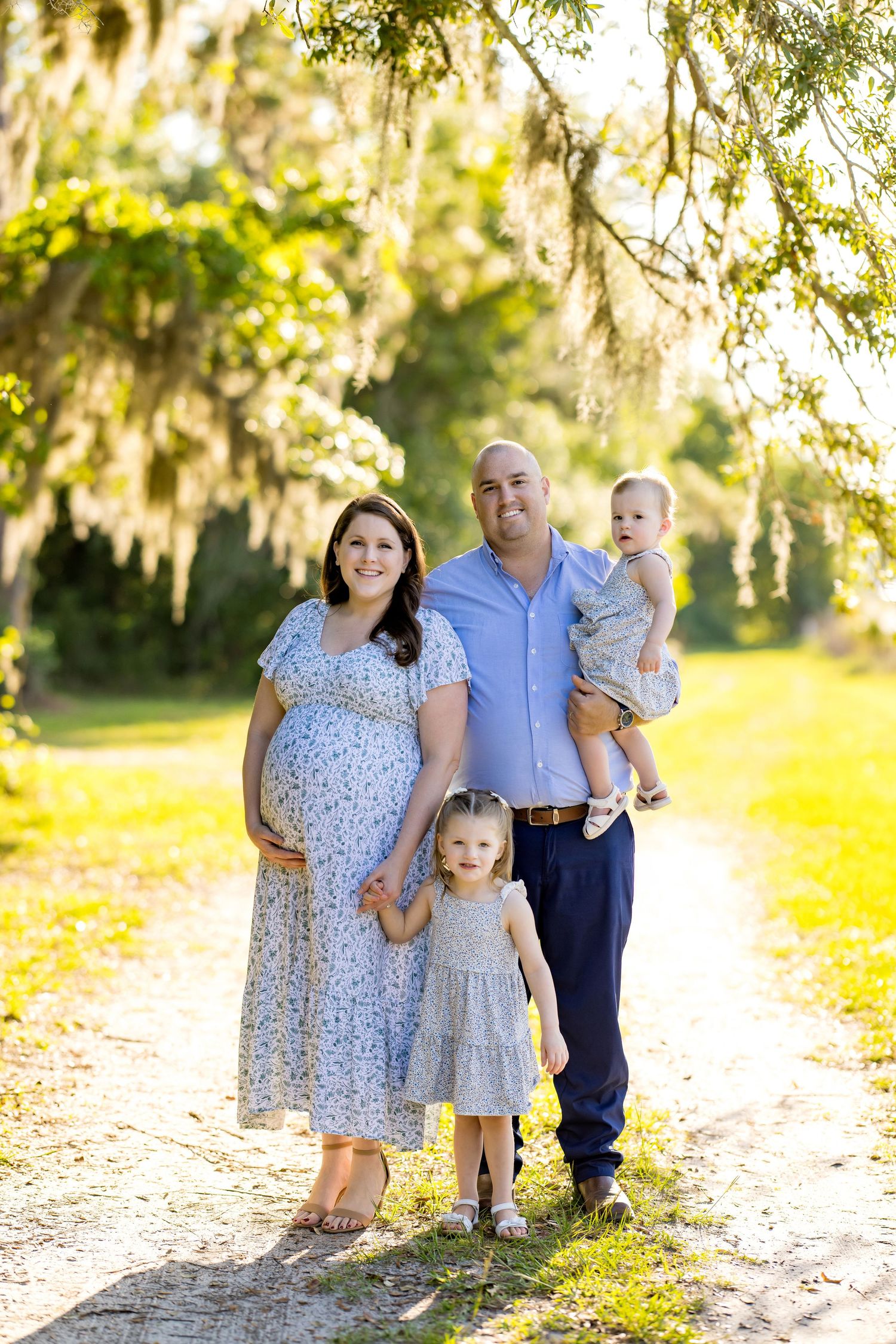 Pregnant woman in floral dress poses with husband and two young children on a sunny tree-lined path.