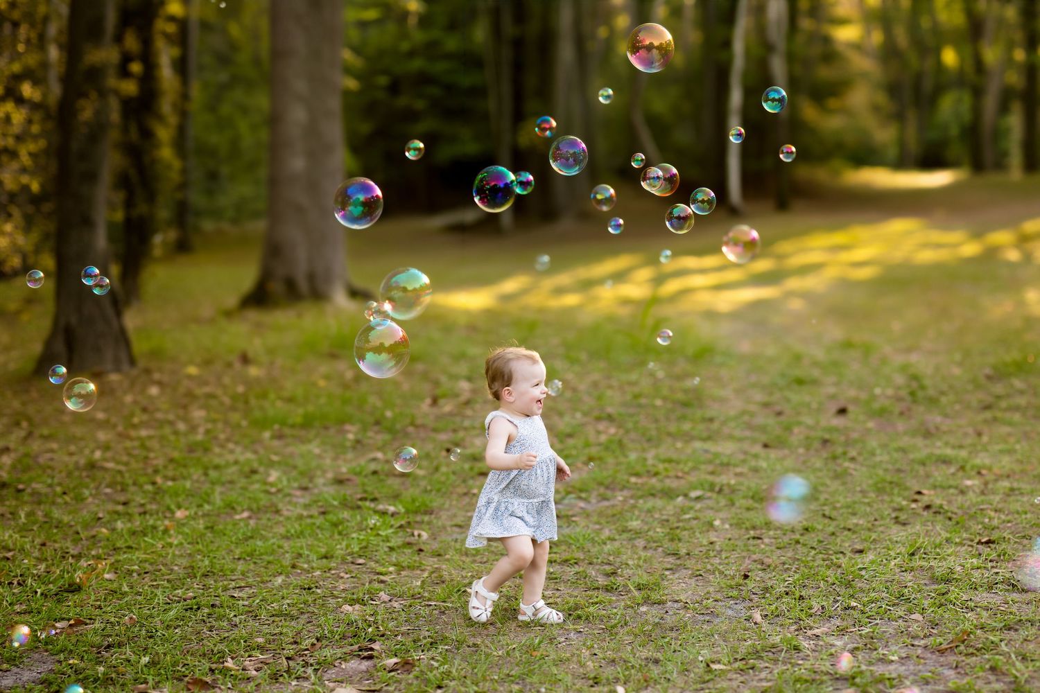 Toddler girl in white dress playing with colorful soap bubbles on a sunny forest path surrounded by green trees.