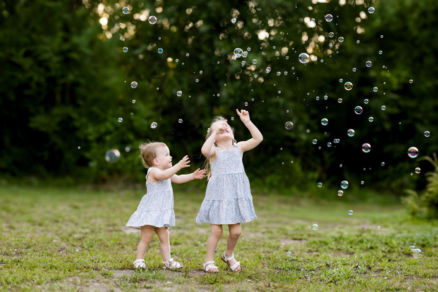 Two young girls in white dresses joyfully playing with soap bubbles outdoors in a green park setting.