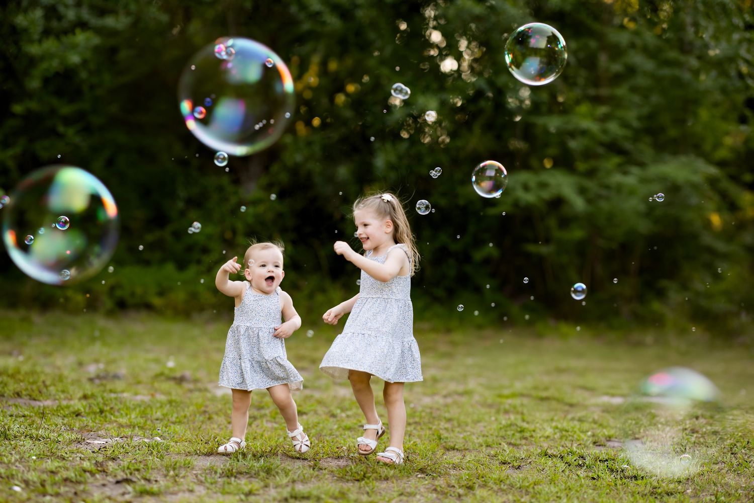 Two young girls in white dresses joyfully chasing soap bubbles in a lush green park on a sunny day.