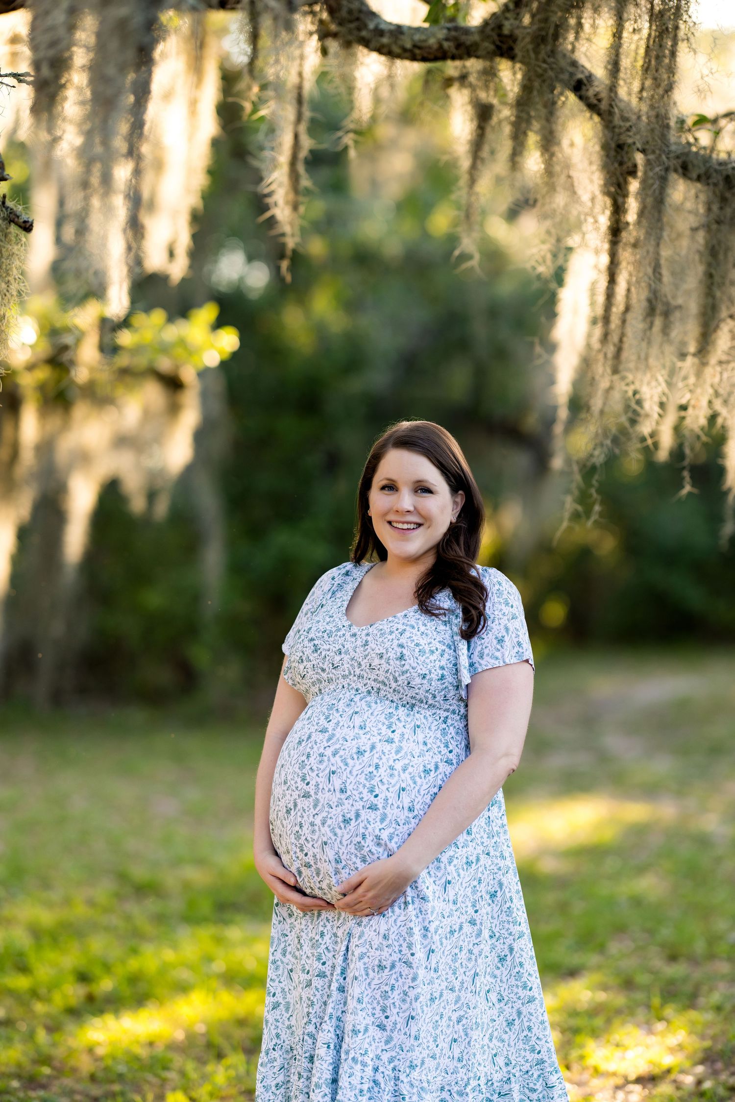 Smiling pregnant woman in blue floral dress cradling baby bump outdoors under Spanish moss-draped oak trees.