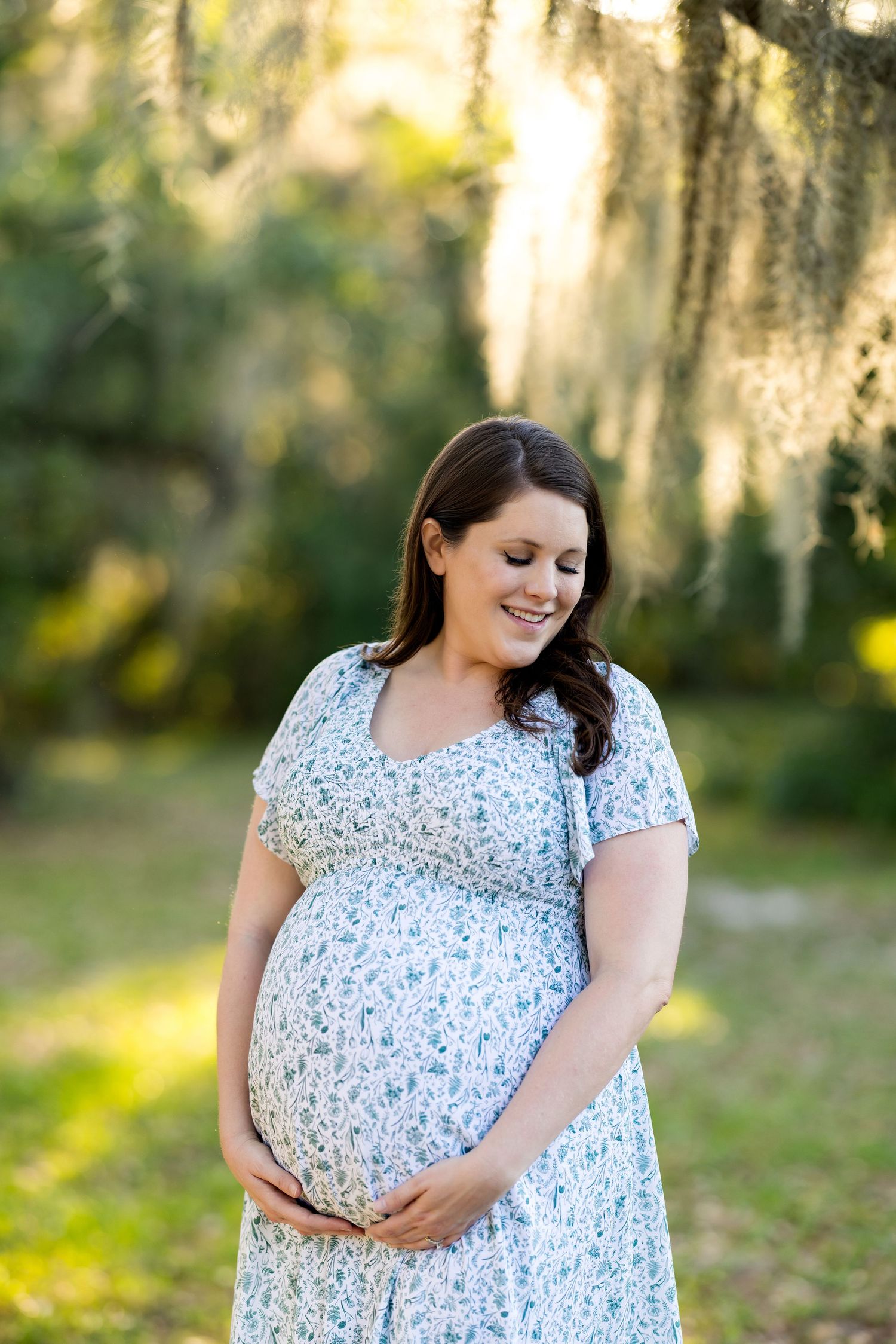 Smiling pregnant woman in floral dress cradling baby bump outdoors under Spanish moss trees.