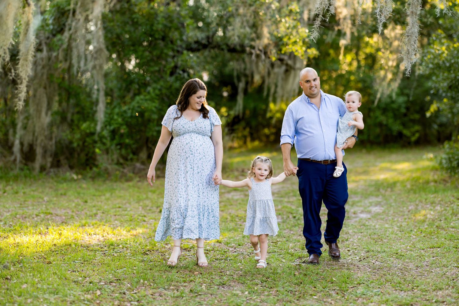 Family walks outdoors with pregnant mother, father holding baby, and young daughter between them.