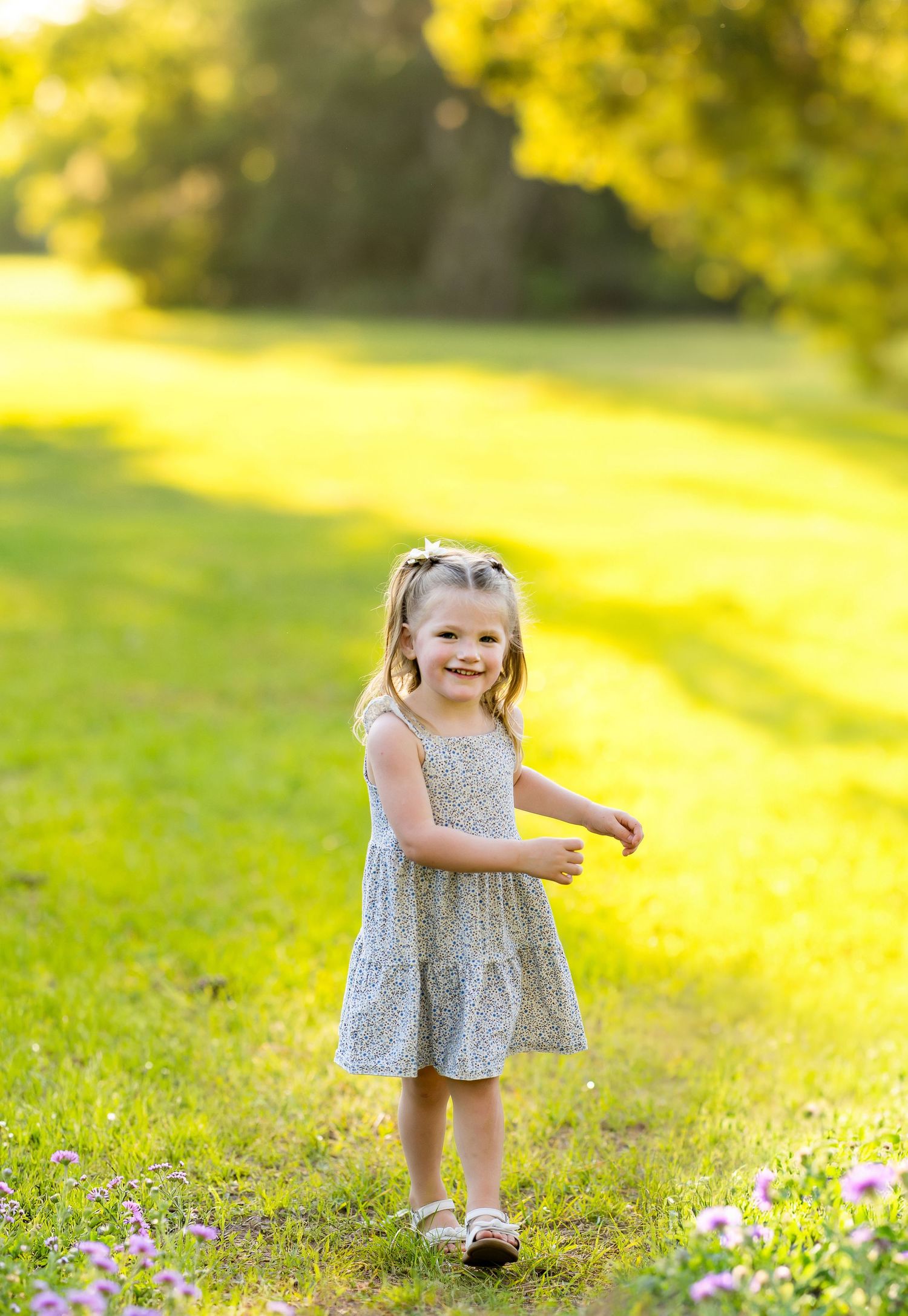 Happy toddler girl in gray dress playing on sunny green grass path outdoors.