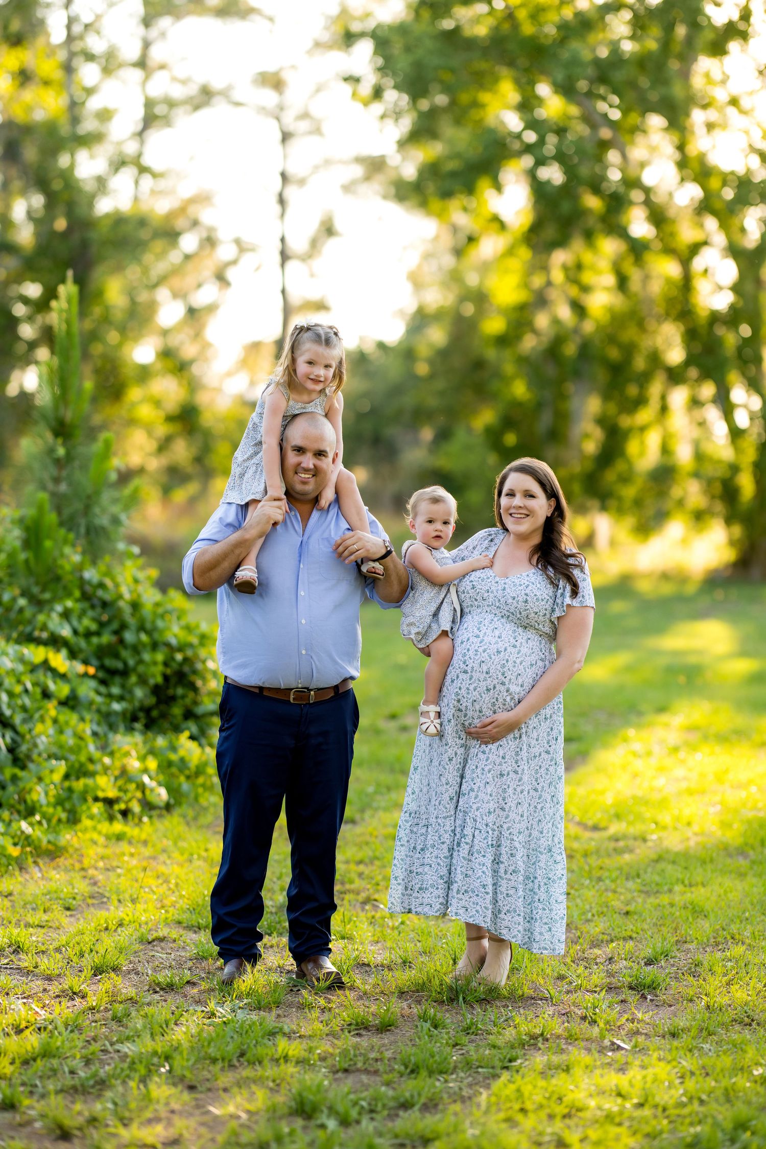 Pregnant woman in floral dress poses with husband and two young children in a sunny green outdoor setting.