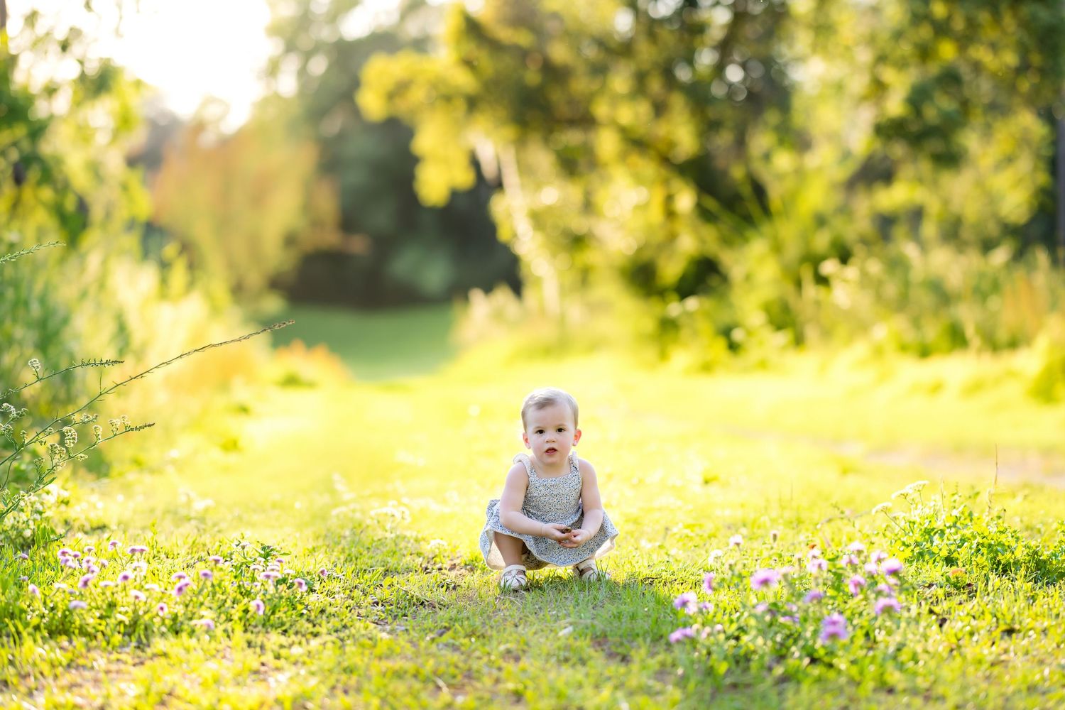A happy baby sits on a sunlit grass path surrounded by wildflowers in a lush green park.