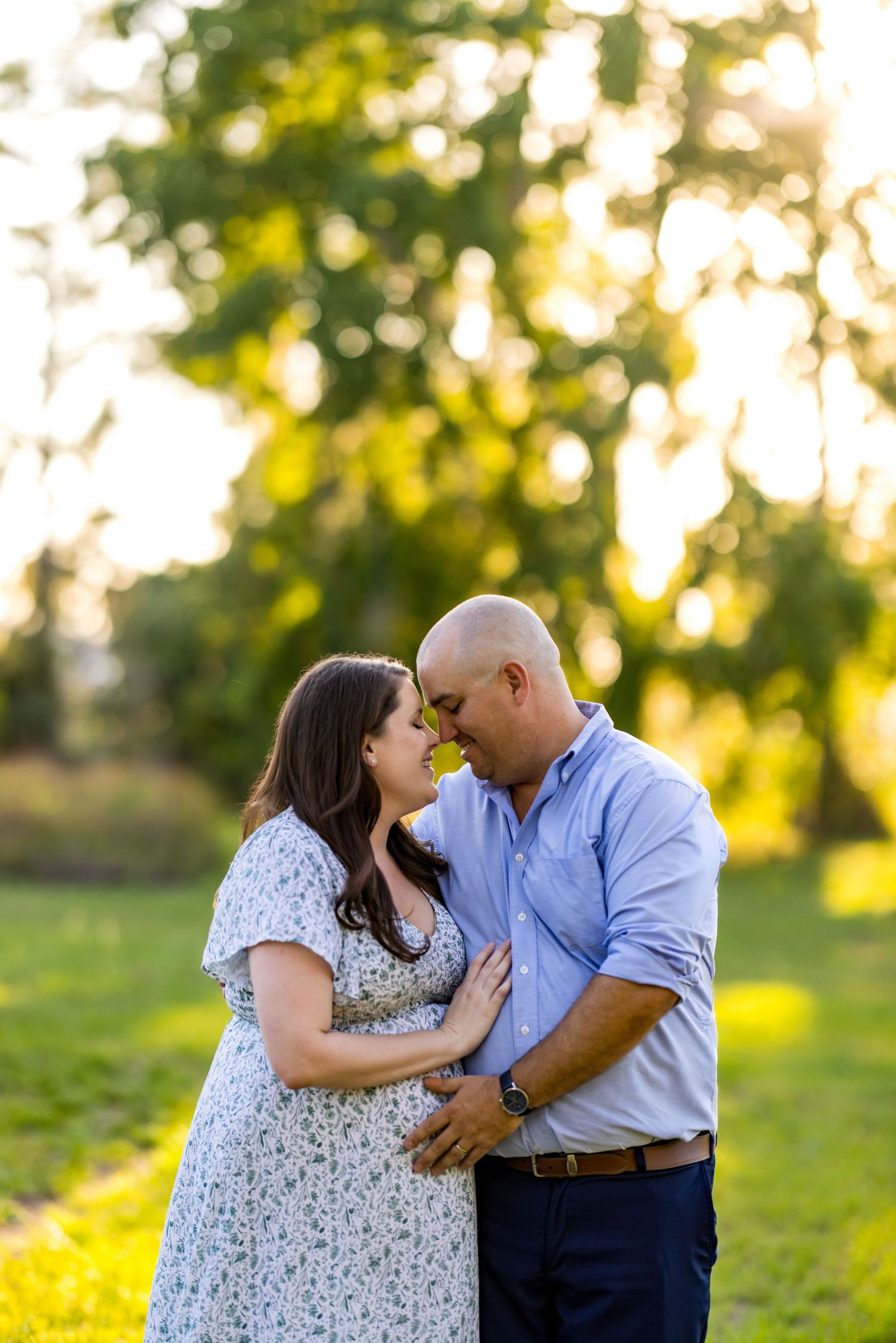 Couple sharing a tender forehead touch outdoors, woman in floral dress with baby bump, man in blue shirt, golden hour.