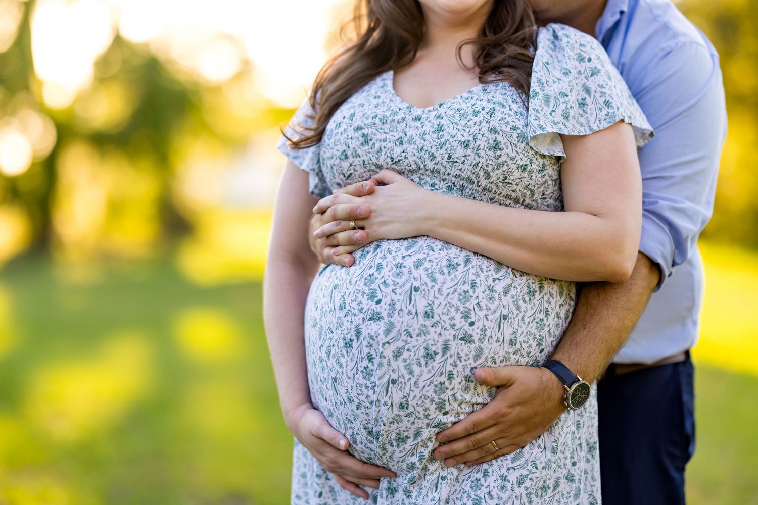 Couple embracing outdoors, pregnant woman in floral dress cradling baby bump in sunny park setting.