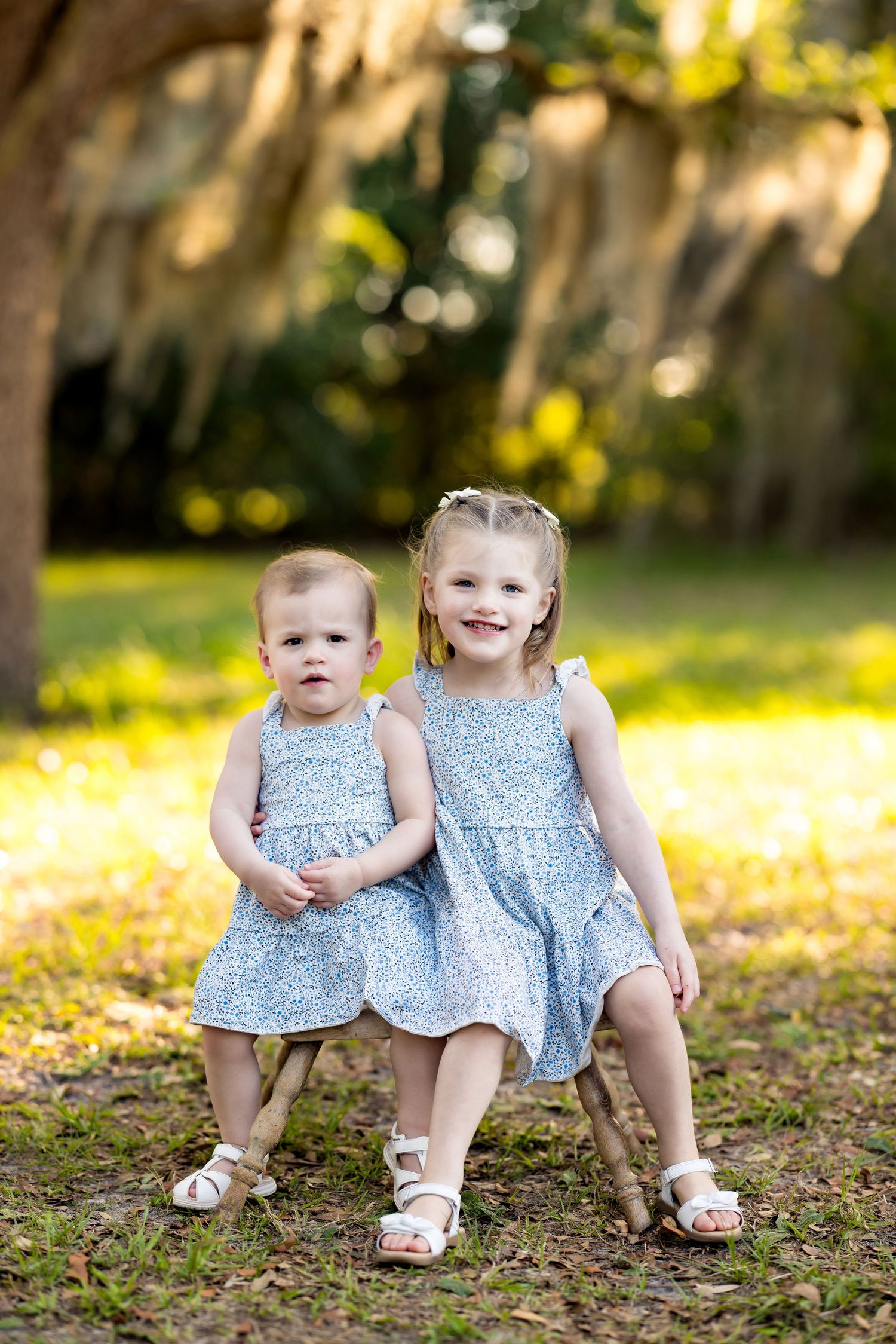 Two young sisters in matching blue floral dresses pose together outdoors under moss-draped trees in golden sunlight.