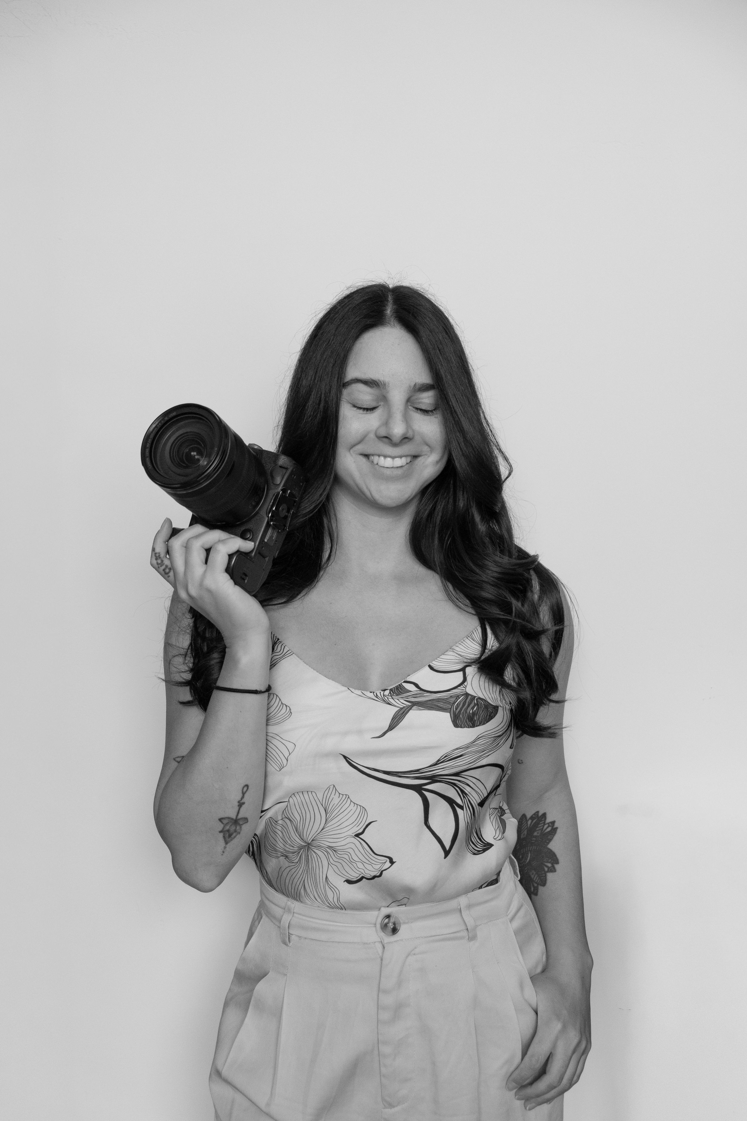 Young woman photographer smiling while holding a DSLR camera, wearing beige outfit, seated against white background.