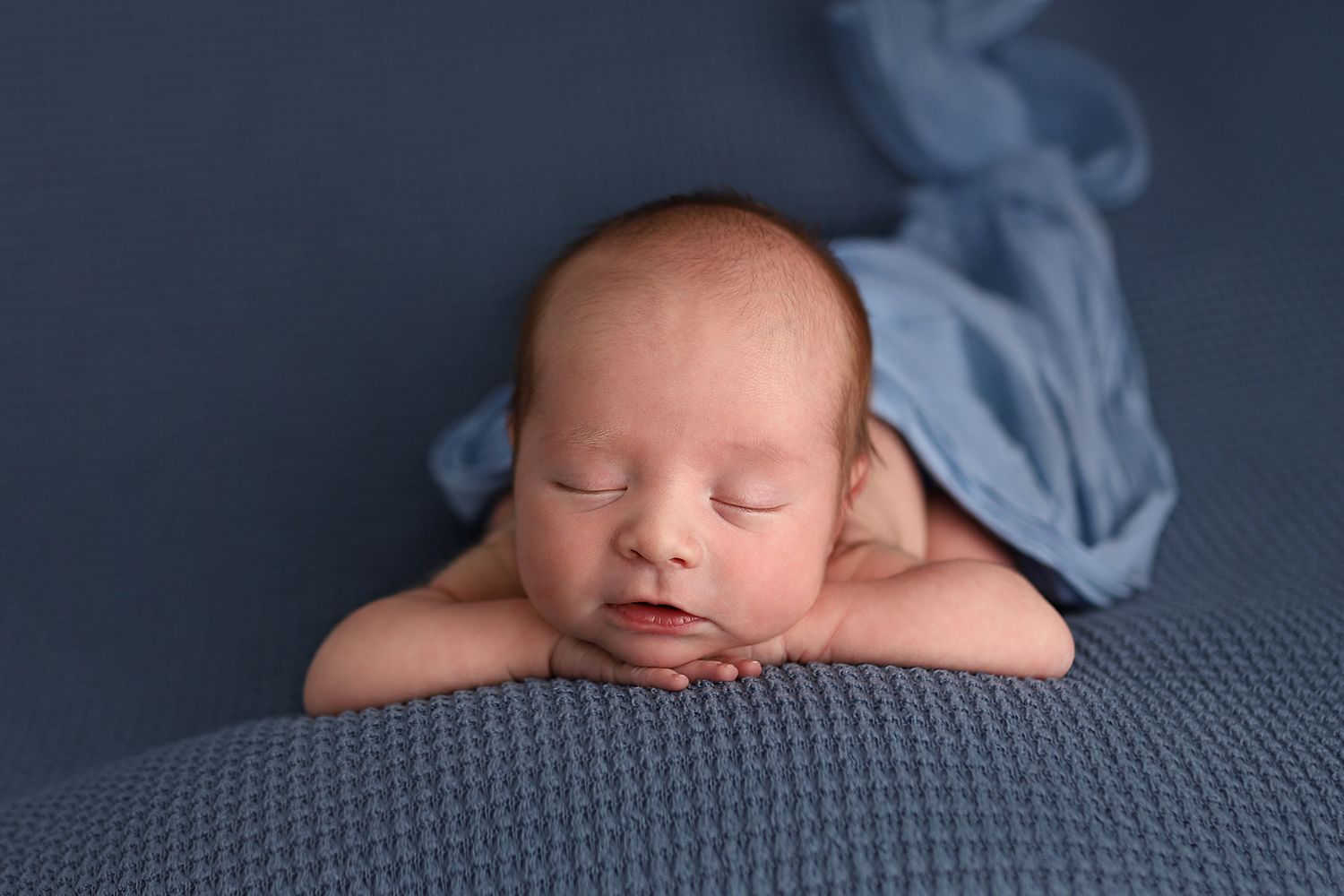 Peaceful newborn baby sleeping on blue knit blanket with hands tucked under chin, wrapped in soft blue fabric.