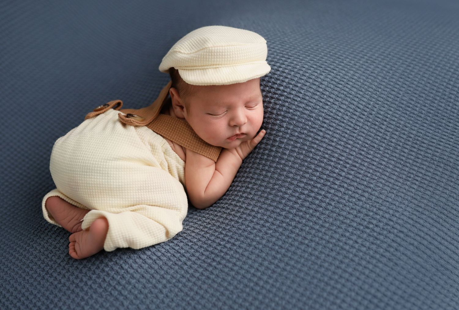Newborn baby in cream outfit and flat cap sleeping curled up on a blue textured backdrop. Utah County newborn photography