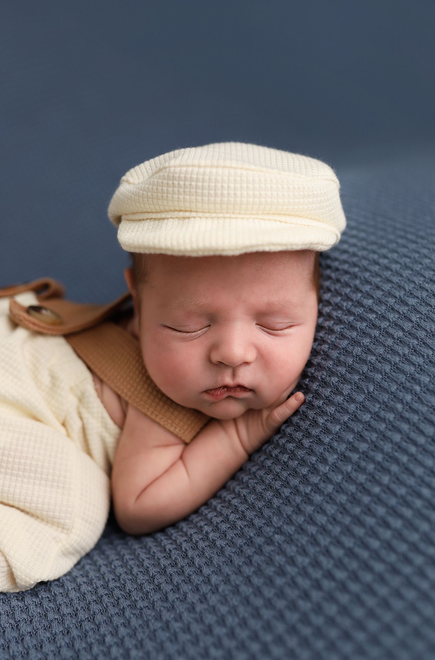 Sleeping newborn baby wearing a cream flat cap hat resting on a blue textured blanket.